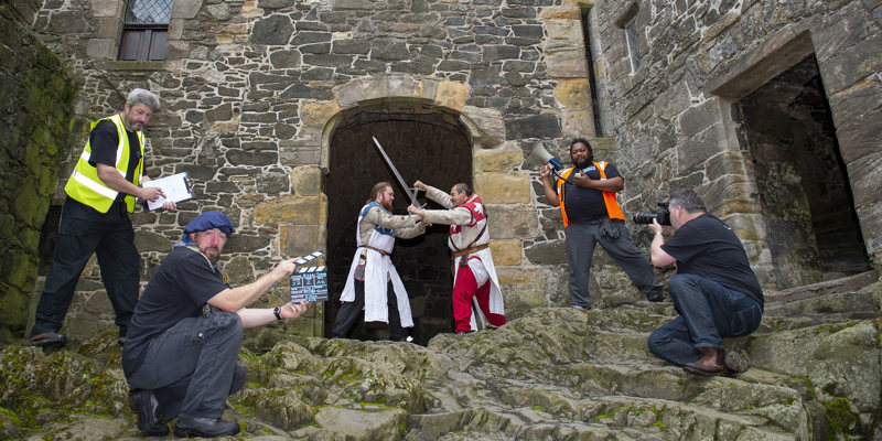 Two re-enactors in the grounds of a castle battling with swords and a crew of 4 with cameras, videos and clapper board