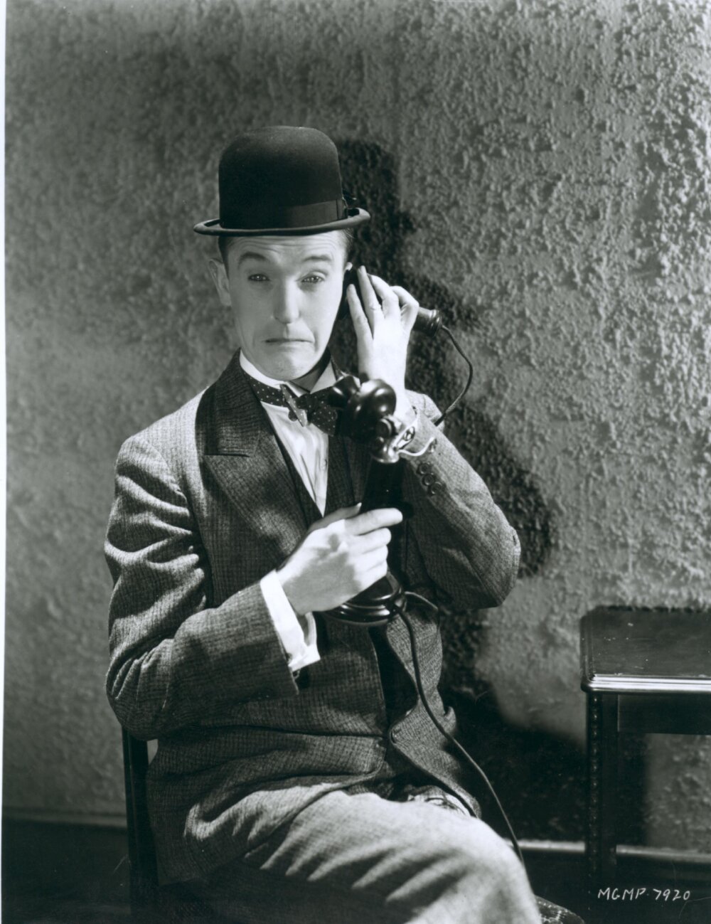Black and white portrait photo of a person wearing a suit and bowler hat, pretending to talk on an old fashioned telephone. They have a sad or confused looking expression on their face.