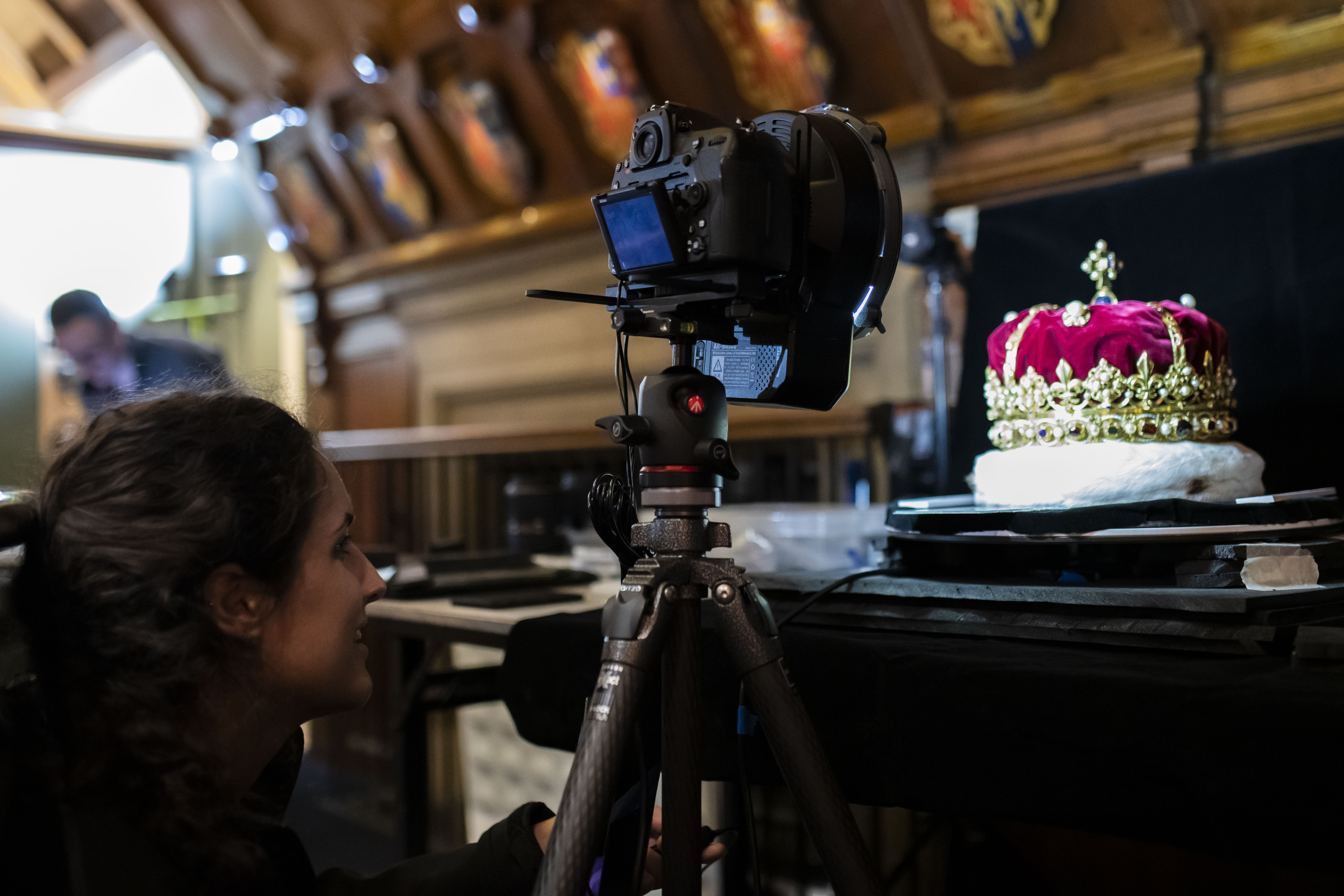 A camera mounted on a tripod photographs a jewel-encrusted crown displayed on a black platform inside an ornate historic room. In the foreground, a person crouches beside the camera, while wood-paneled walls and decorative shields are softly blurred in the background. The scene is warmly lit, highlighting the crown and the imaging process.