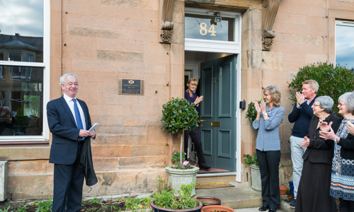 A plaque is installed by the entrance to a building. People stand in front clapping.