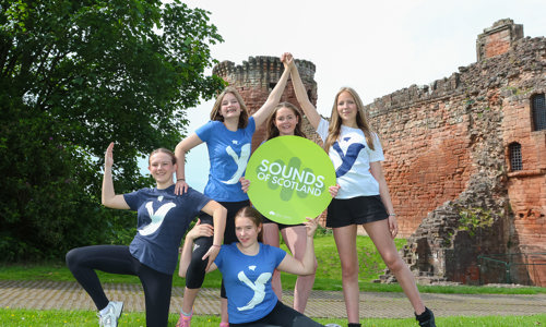 Five teenage girls doing dance poses, standing together with one sitting on the grass in front. Three girls have blue t-shirts on while one has white and another has a navy tshirt on. In the middle, one of the girls is holding a green board in the shape of a circle with white writing on it which says Sounds of Scotland. A red stone castle is in the background.