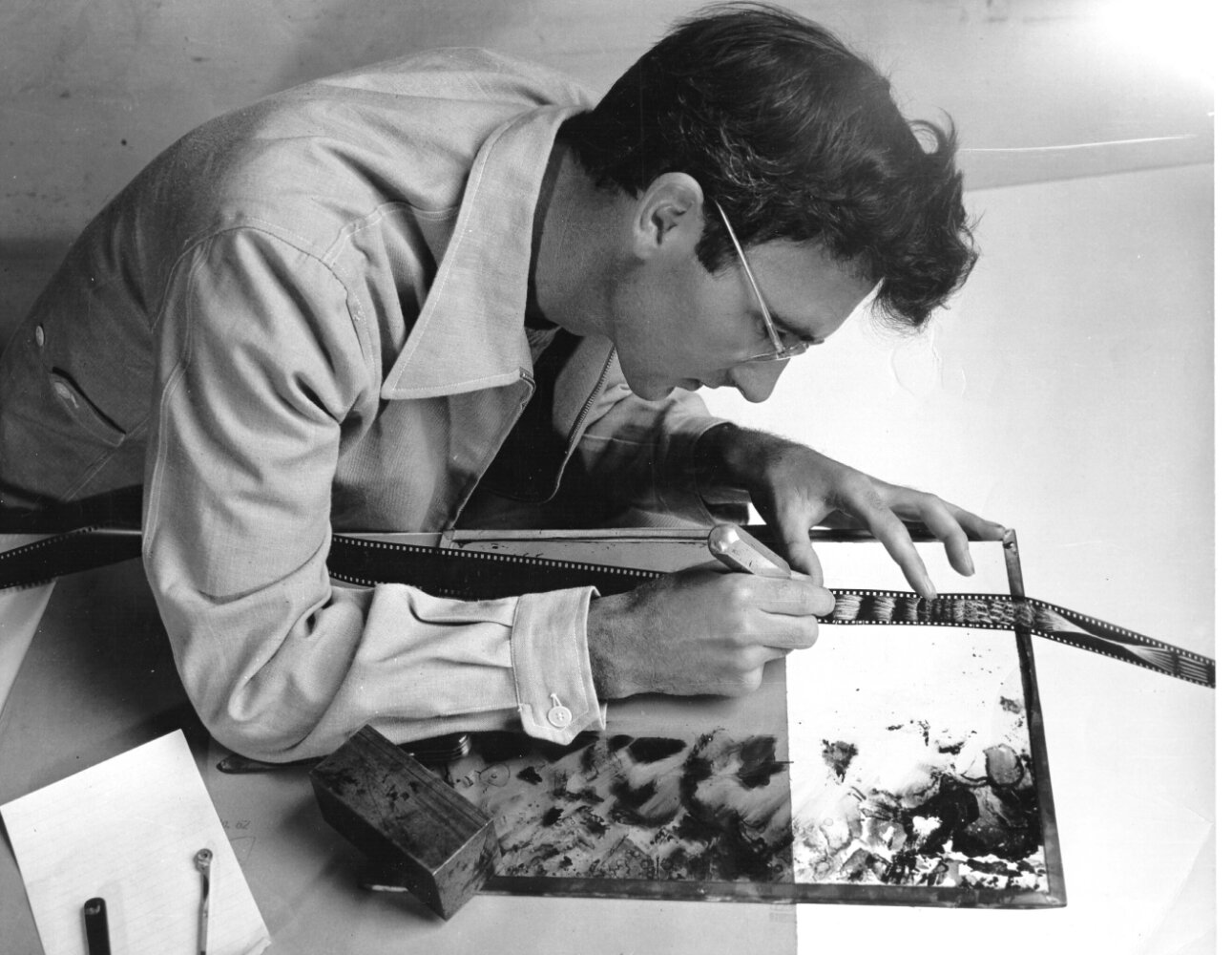 Black and white photo of a person in a shirt and glasses leaning over a table. They are marking a series of film negatives with a pen, as light shines upwards from underneath.