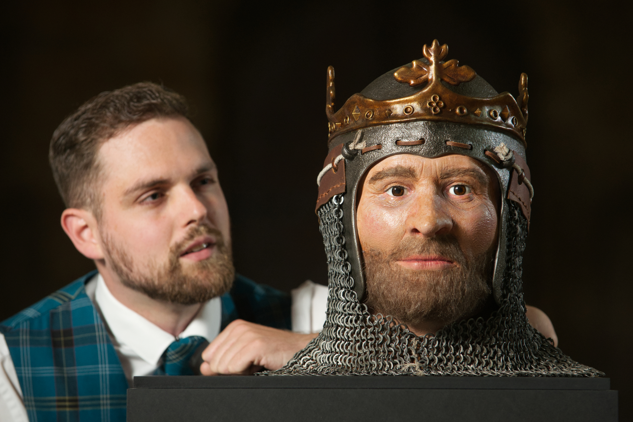 A man stands beside a detailed historical reconstruction of Robert the Bruce's head, face and shoulders. Bruce has a short beard, and wears a medieval helmet with a decorative crown and chainmail/.