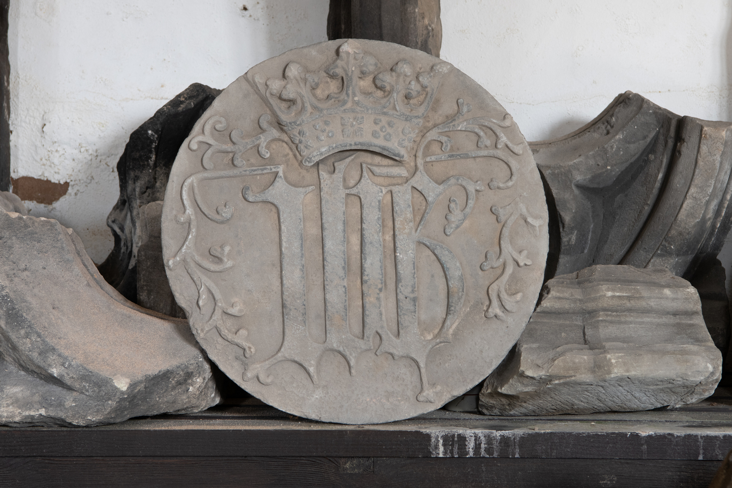 A flat, circular stone artefact, with markings on it. There is a crown at the top, what looks like two letters underneath and leaf-like patterns around the edges. There are broken stones surrounding it. 