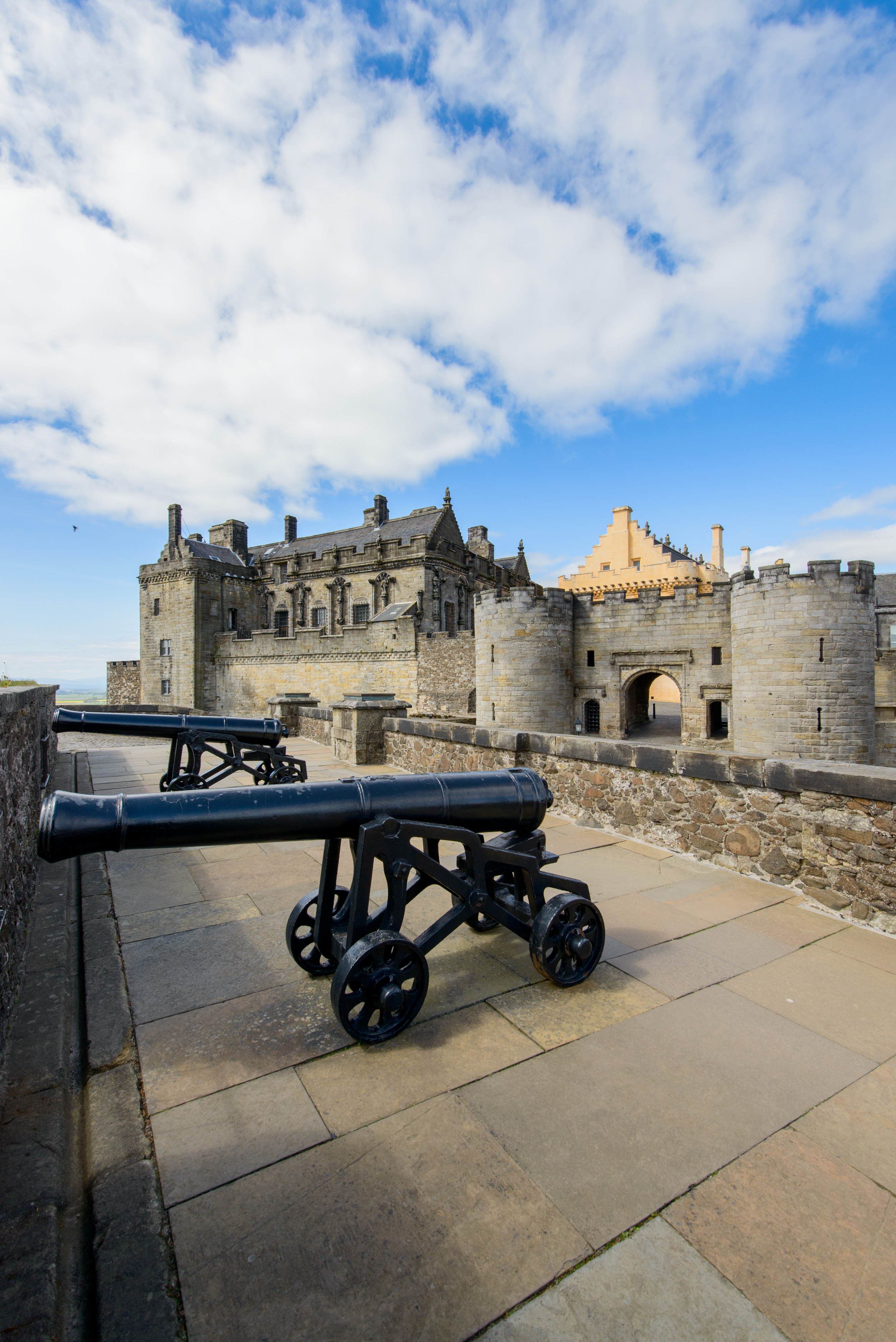 Historic castle with several different stone buildings with shiny black cannons in the foreground under a blue sky with white clouds.