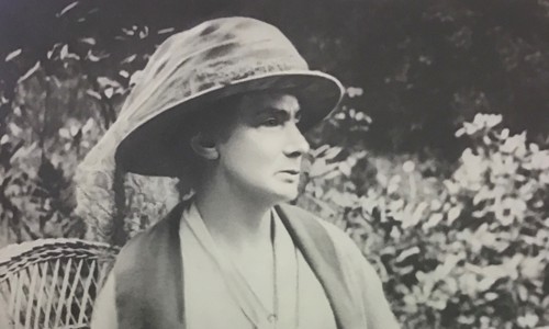 Black and white image of woman sat in wicker chair in a garden. She is wearing a hat and holding a handbag on her lap.