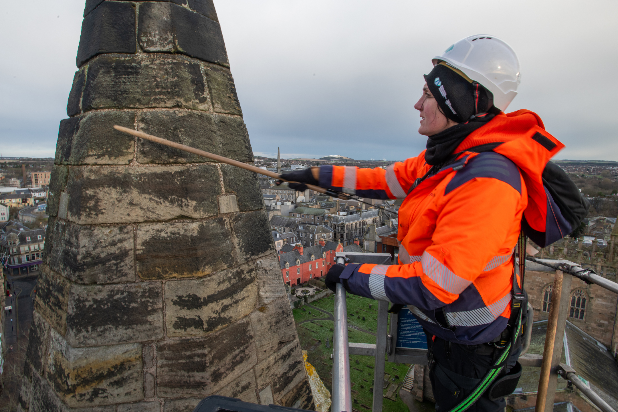A worker in high-visibility protective gear stands in a raised platform, examining the stone surface of an abbey tower with a long pointing tool.