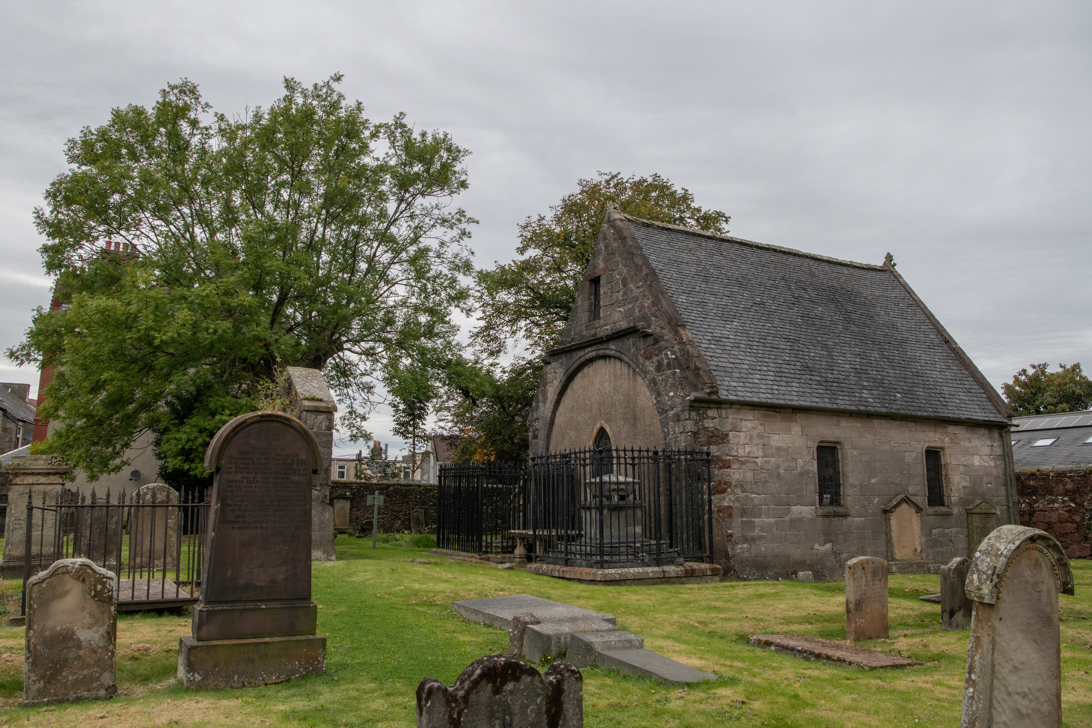 Exterior view of the Skelmorlie Aisle within the cemetery, showing the stone building with a pitched slate roof, surrounding gravestones, and a large leafy tree.
