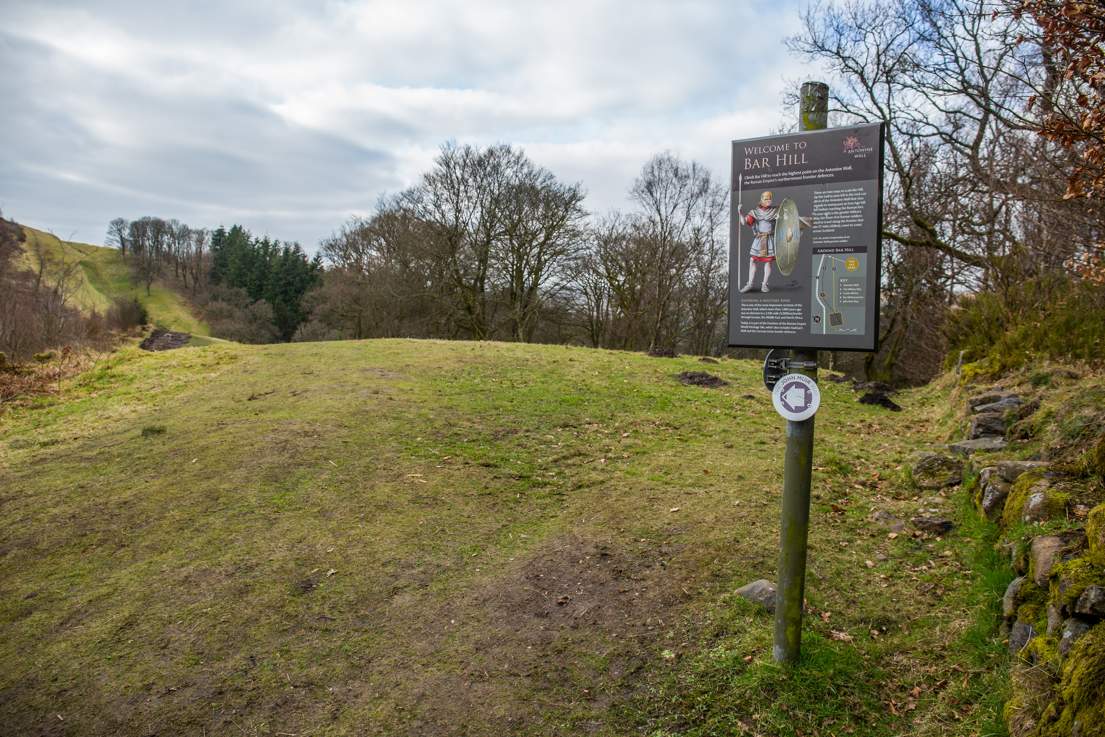 Information sign at Bar Hill Fort mounted on a wooden post, featuring text and an illustration of a Roman soldier holding a shield. The sign provides historical details about the site. It stands on a grassy path with trees and rolling hills in the background under a cloudy sky.