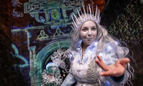 A woman with long silver hair dressed in a white and silver dress with lights is wearing a white crown and holds her hand out to the camera. Behind her, there are blue images projected onto a stone castle wall.