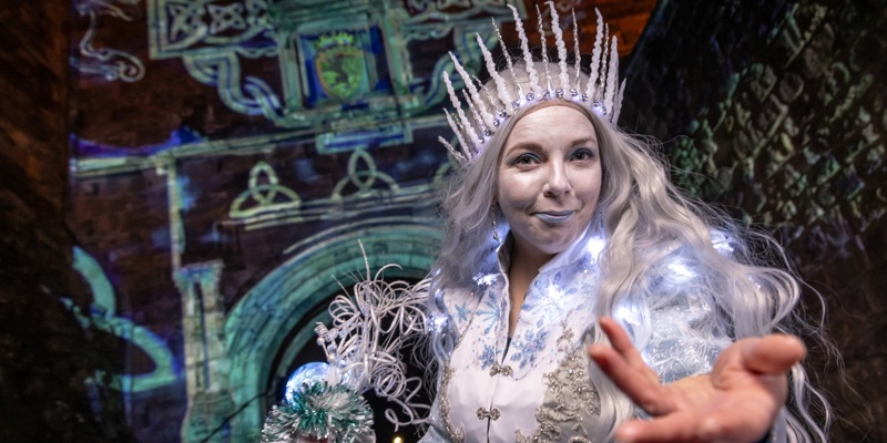 A woman with long silver hair dressed in a white and silver dress with lights is wearing a white crown and holds her hand out to the camera. Behind her, there are blue images projected onto a stone castle wall.
