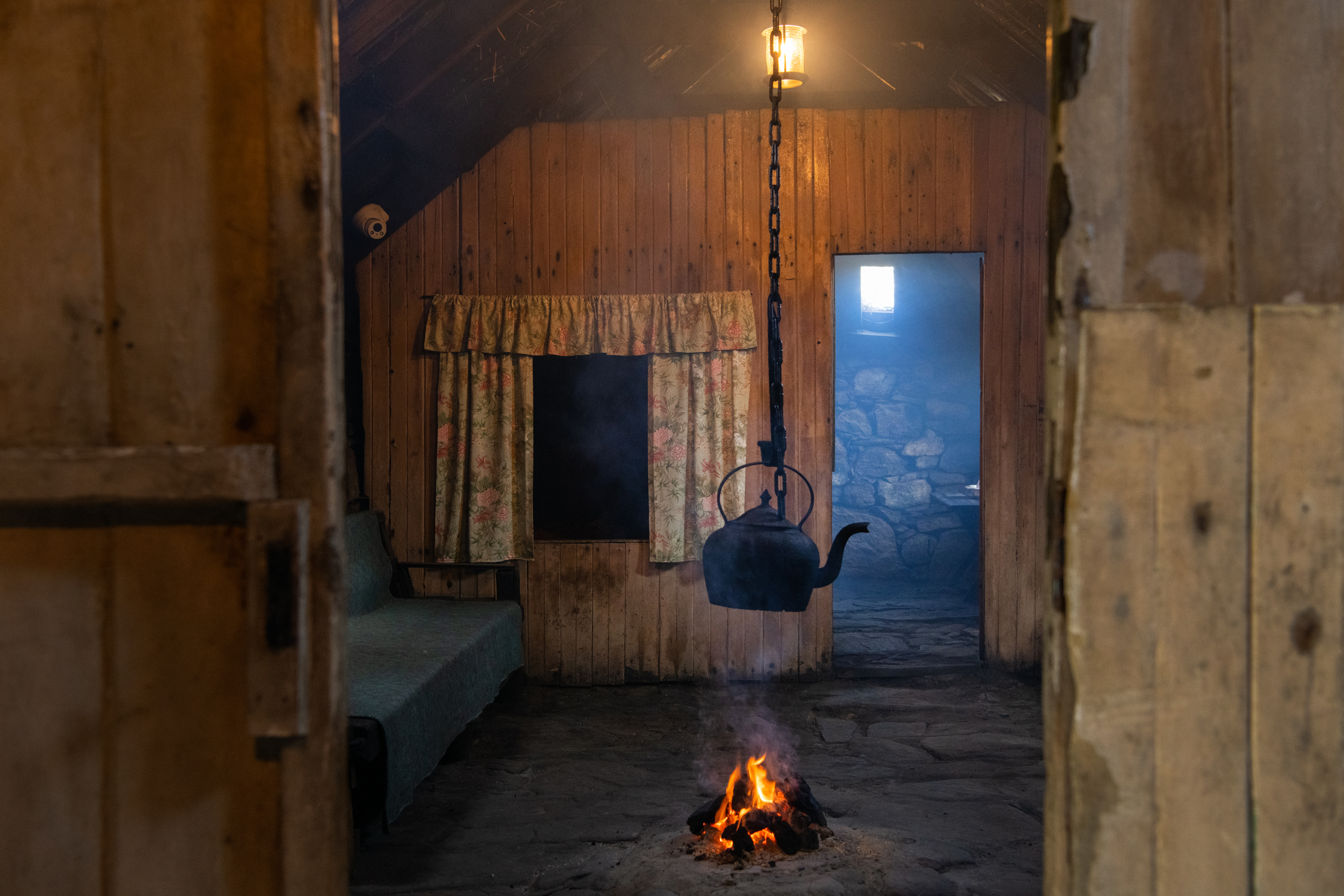 An iron kettle hanging from a chain with a fire burning beneath it, in the middle of a room made with wooden walls