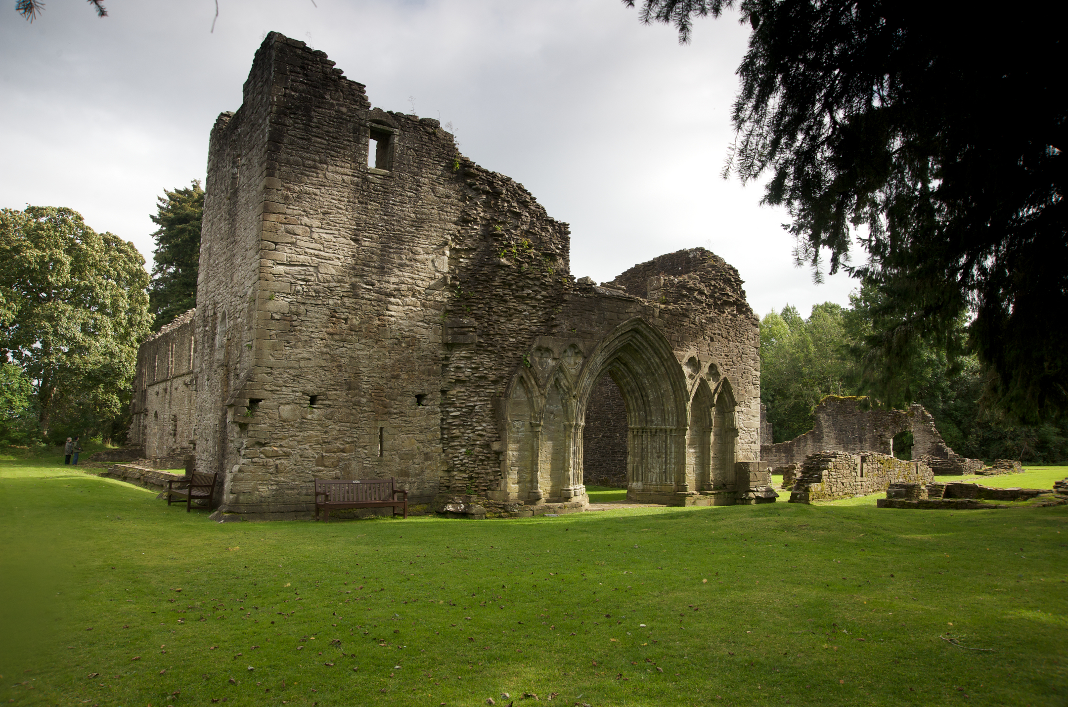 Stone ruins of Inchmahome Priory with large Gothic archways and partial walls, surrounded by grass and trees.