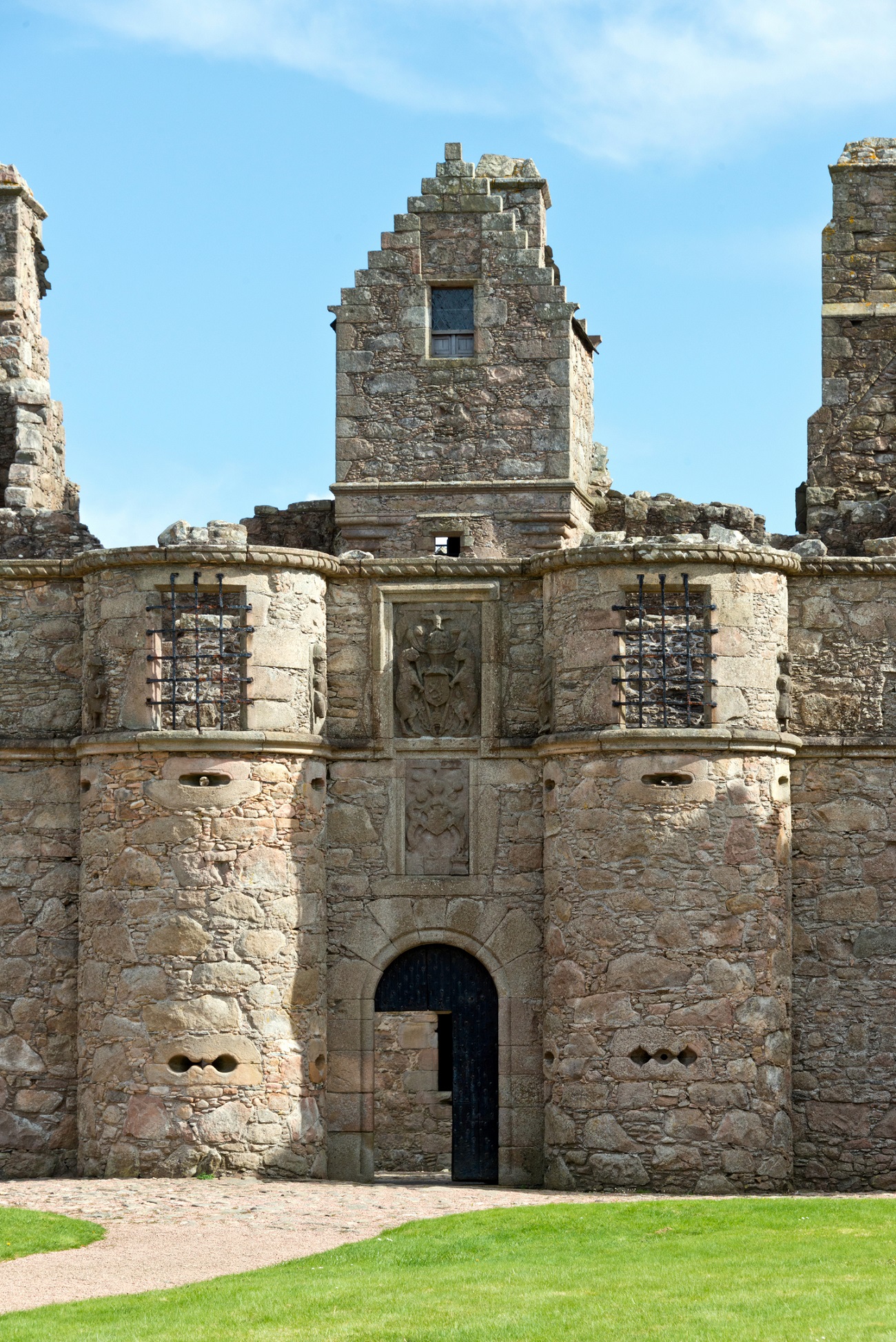 The front entrance of Tolquhon Castle, featuring tall, weathered stone towers and decorative stonework under a bright blue sky.