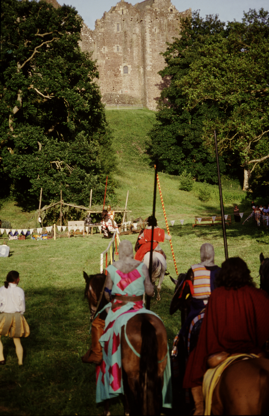 People dressed up in medieval outfits sit on horses, facing a stone castle that sits on a mound. There are large trees either side of the castle. 