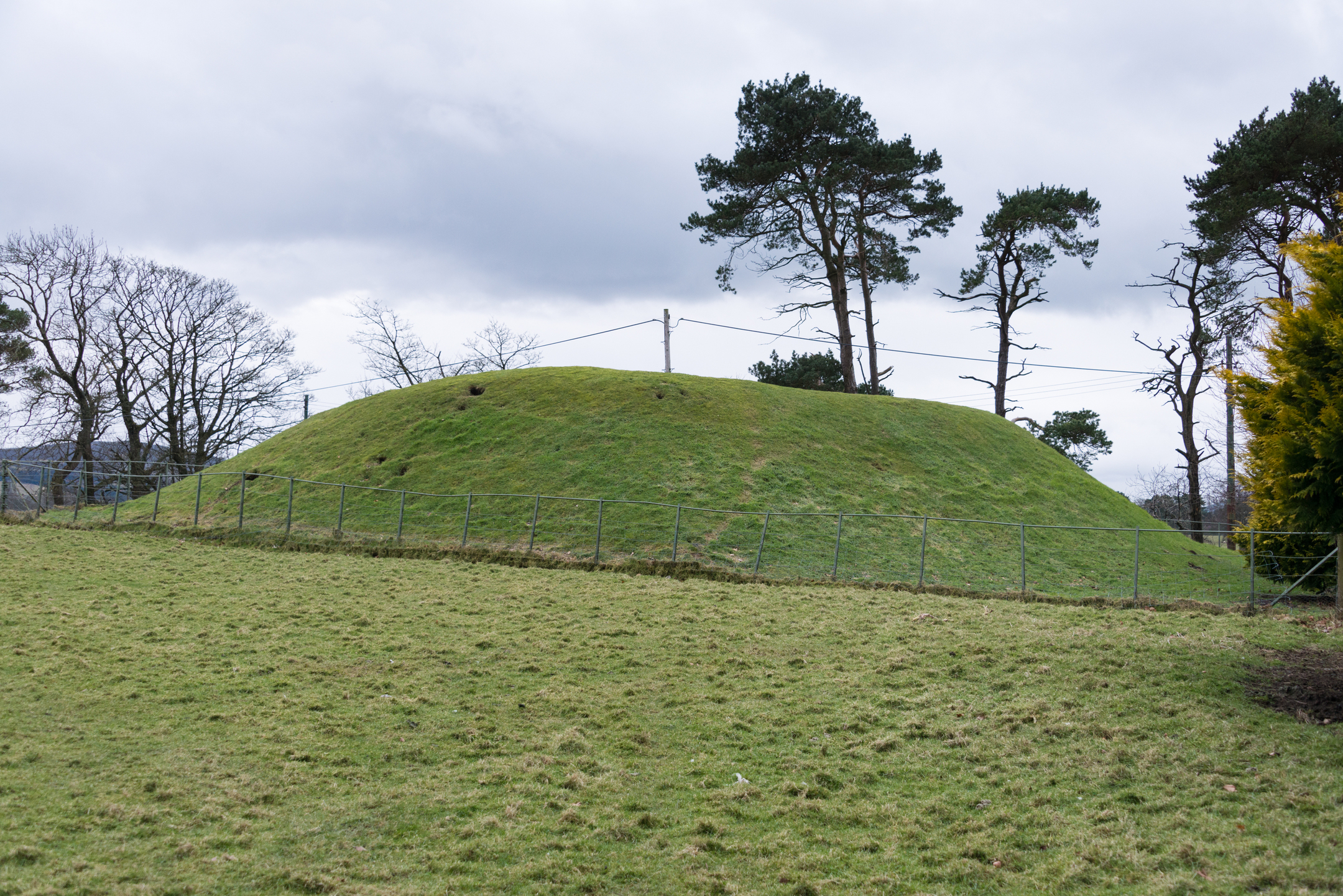 A historic site consisting of a grassy mound which may have once been topped by a settlement or fort. The mound is in a farmer's field and separated from the field by a small metal fence.
