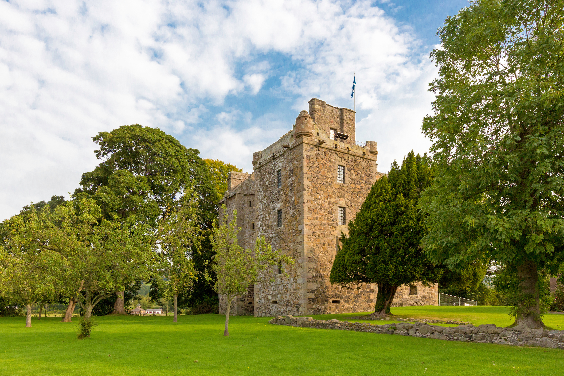 Elcho Castle seen from the garden in summer, with bright green grass and leafy trees surrounding the stone tower house. A Saltire flag flies from the roof against a partly cloudy blue sky.