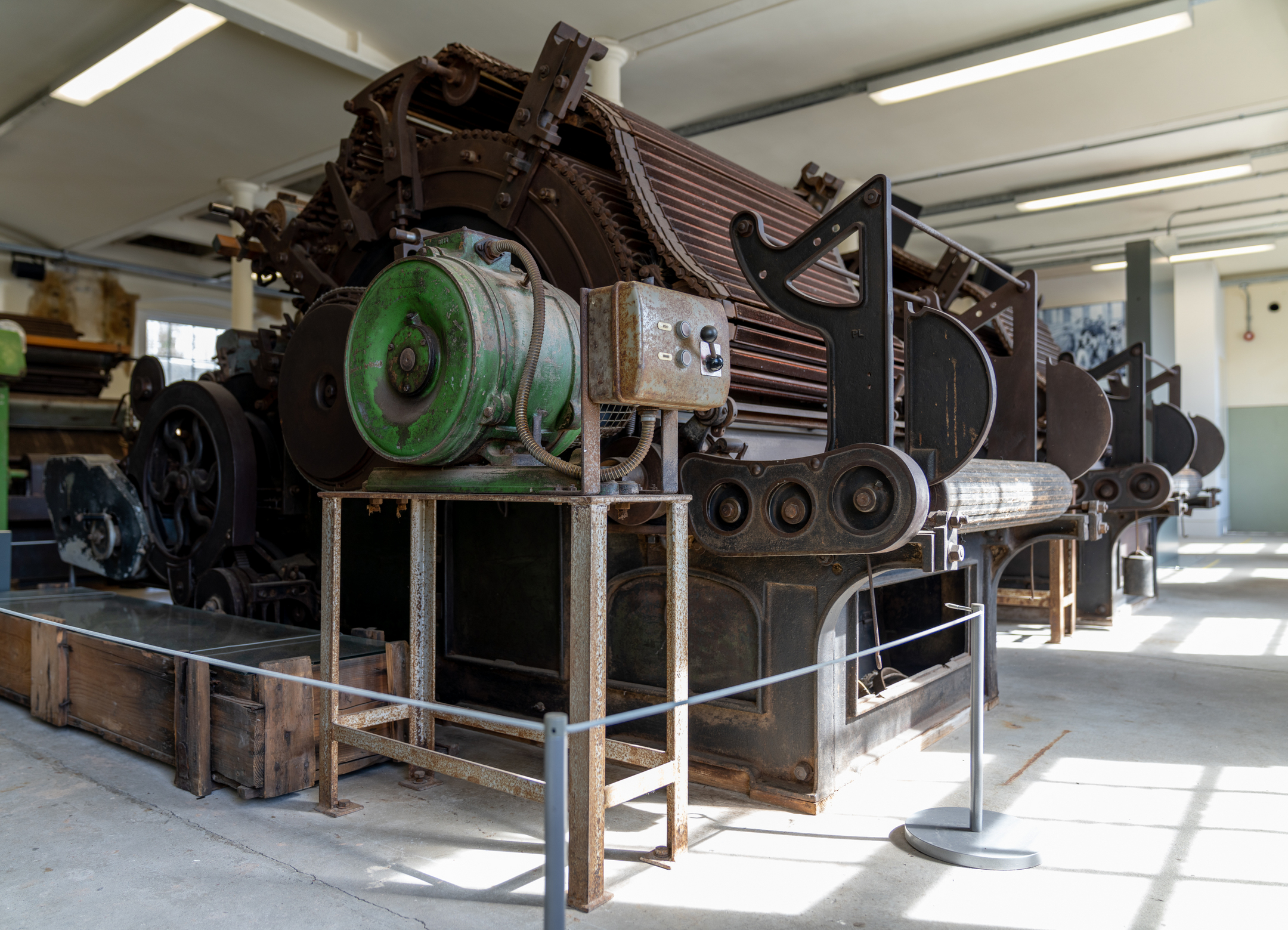 A row of large, rusted industrial machines with gears, rollers, and a green motor displayed inside the mill's expansive workshop area.