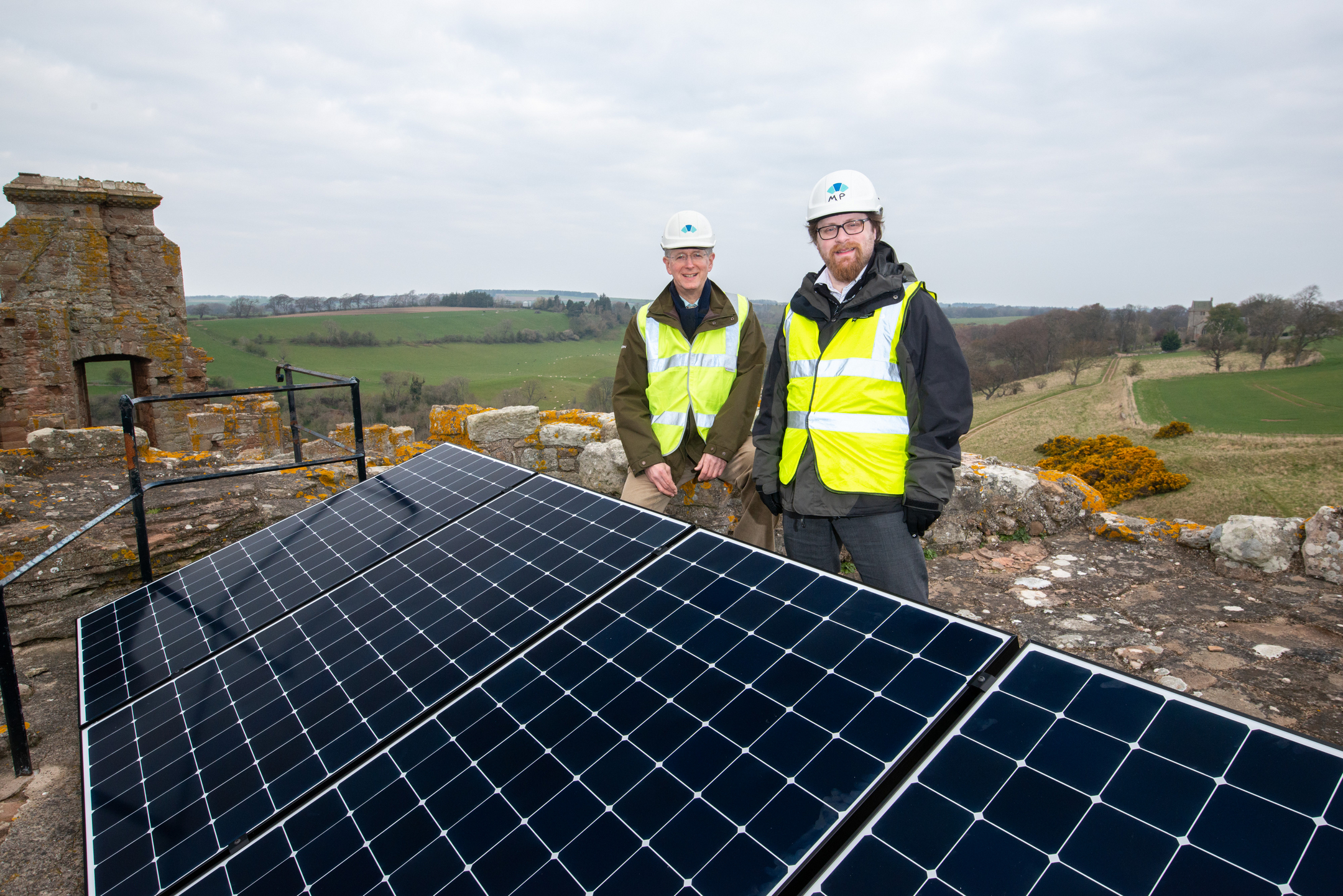 Two men wearing hard hats and hi-vis jackets stand on the roof of a medieval stone castle behind four solar panels. It is a cloudy day.