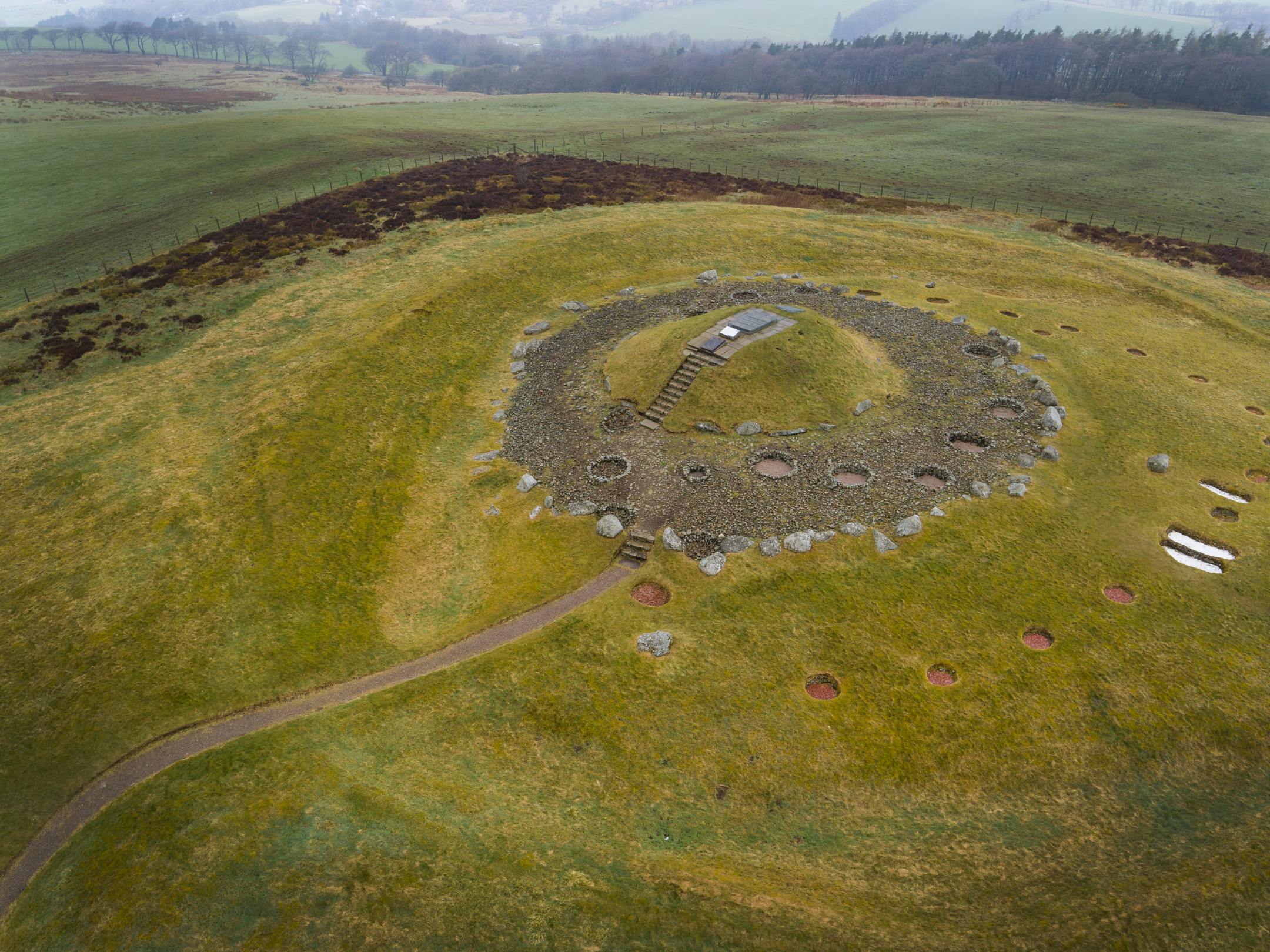 Aerial view of grassy area with a path leading to a circular area with a mound in the middle and stairs leading up to an entrance. There are stones and intentional holes dug in the area too