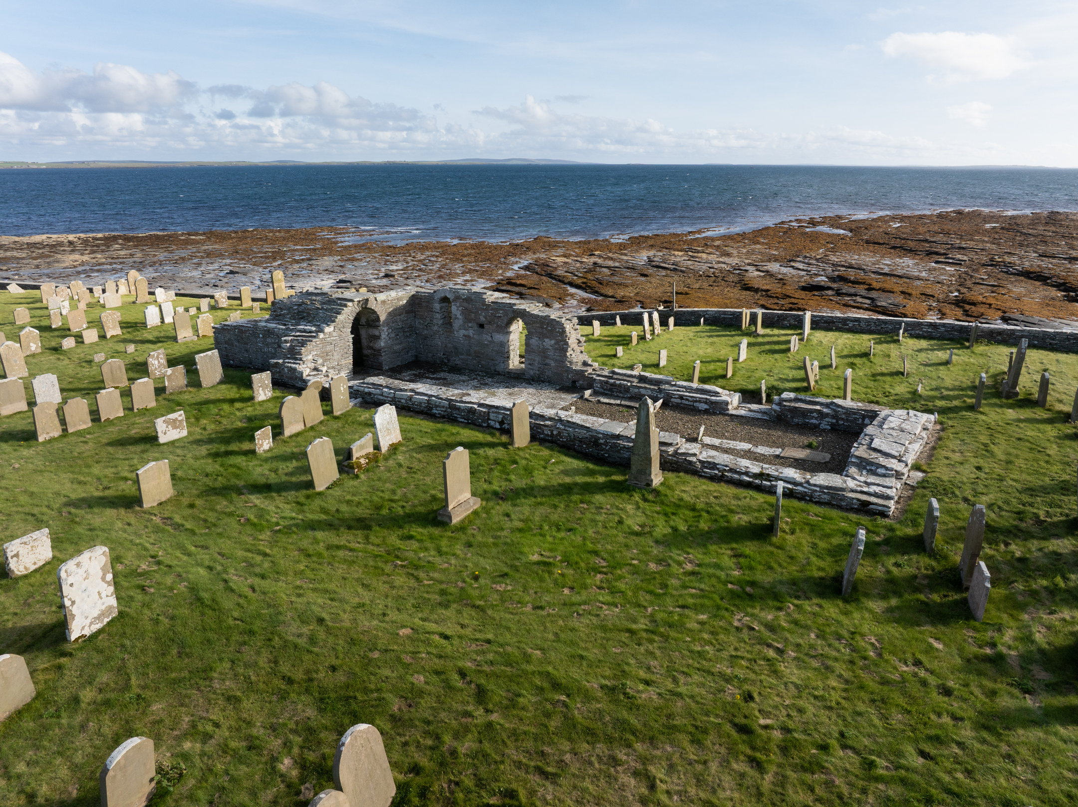 Aerial view of the entire church ruin and surrounding graveyard, set on a green slope above the rocky shore and looking out over the sea.