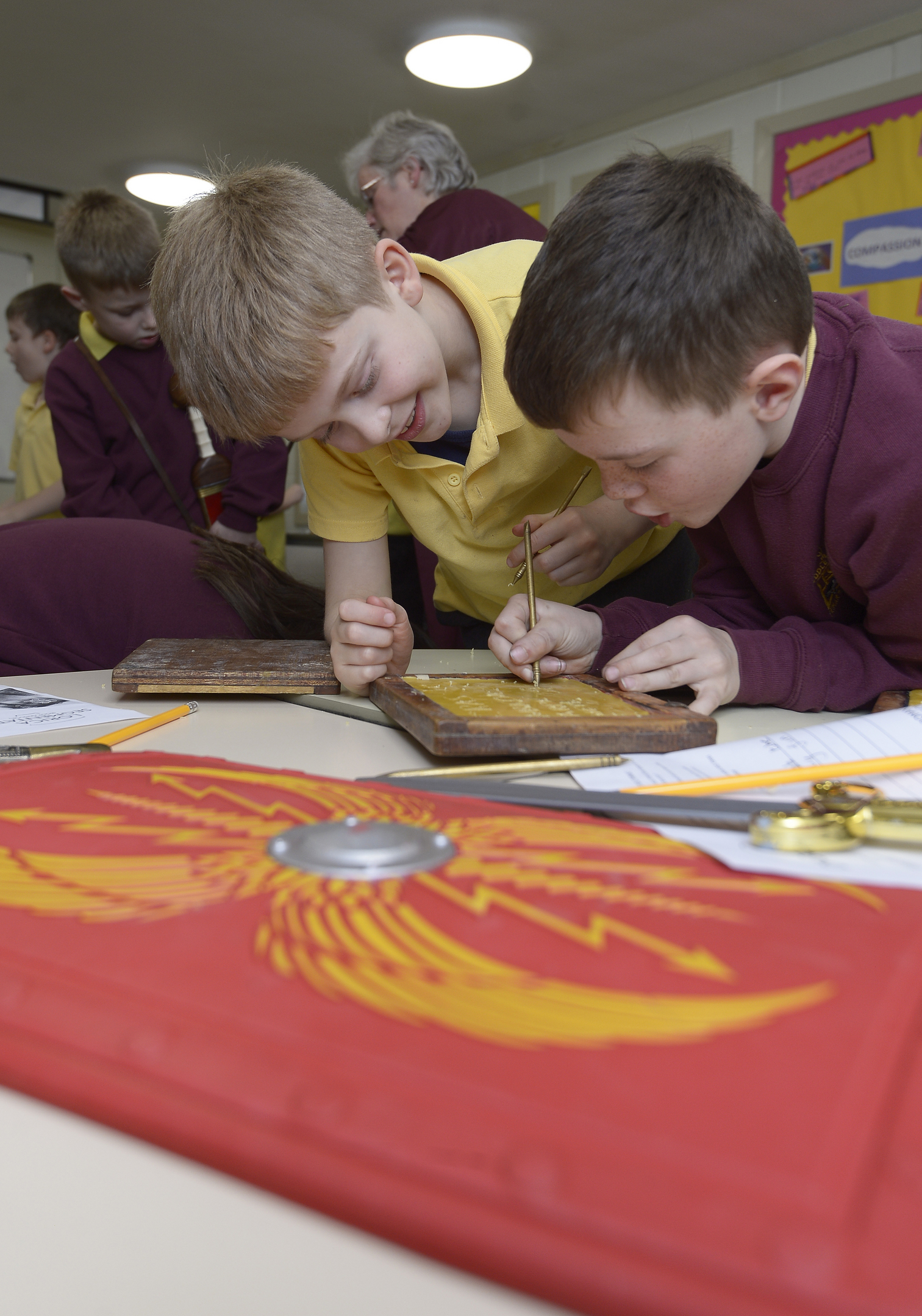 Two school children sat at a table writing on a replica Roman wax tablet with a replica shield next to them