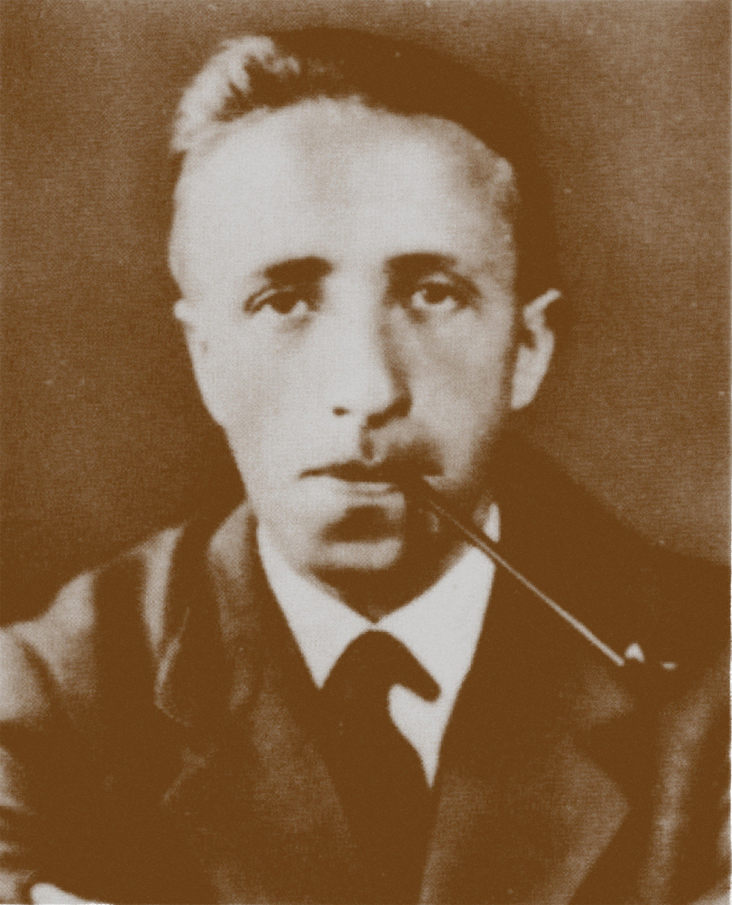Sepia-toned, slightly soft-focus portrait of a person from the early 20th century, shown from the chest up. The person wears a dark suit jacket, white shirt, and dark tie, and holds a pipe at the mouth. The hair is short and swept back, and the background is plain and evenly lit.