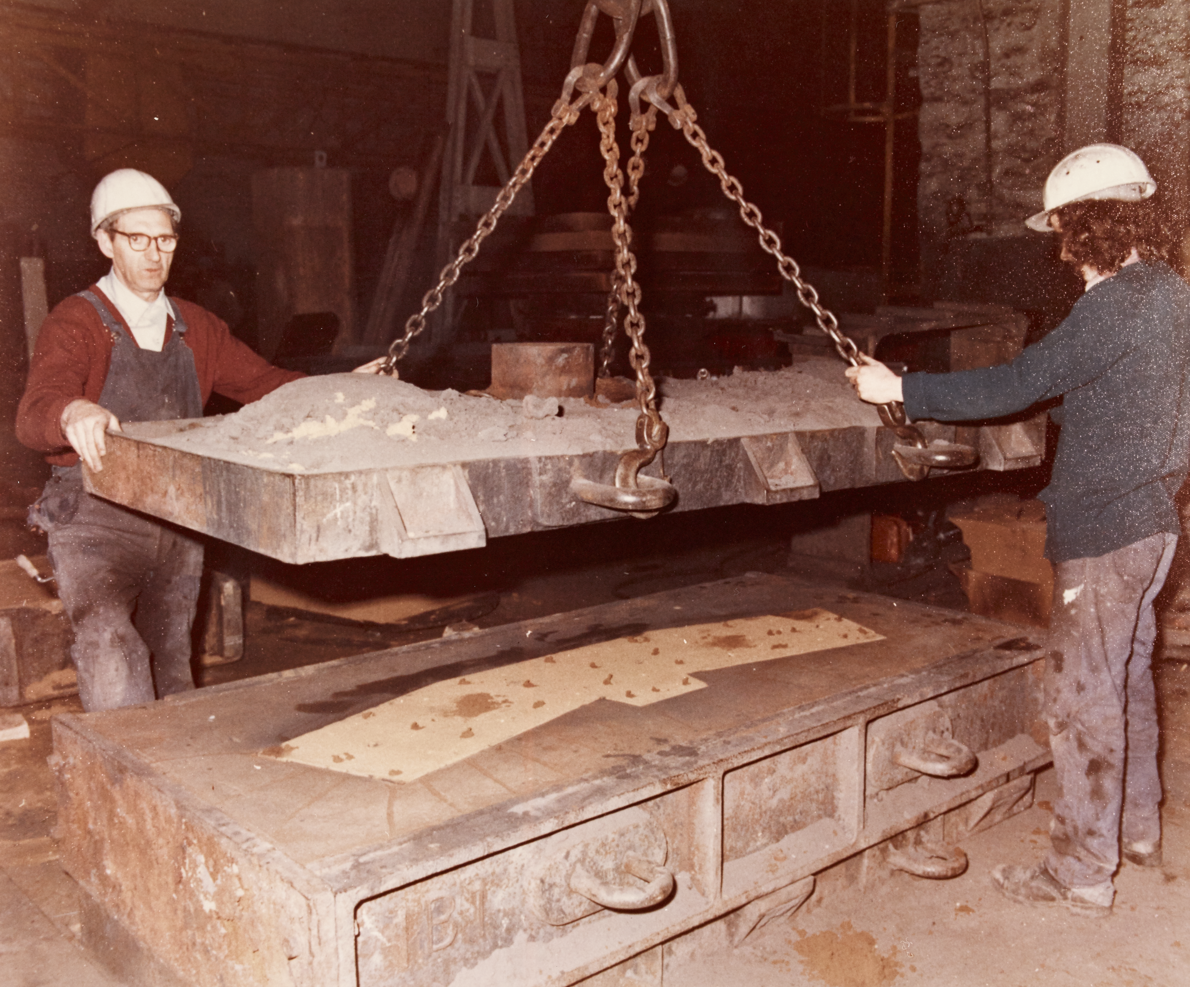 Equipment being moved, with a heavy item held up by chains and two men in safety hats