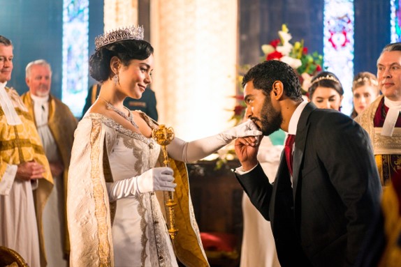 A woman in an ornate white gown and crown stands in a church, smiling as a man in a dark suit bows to kiss her hand during a formal ceremony.