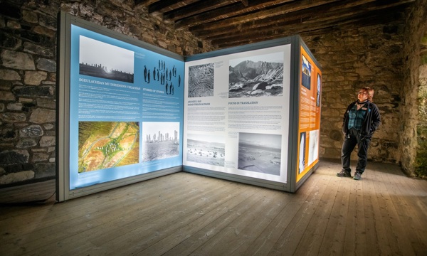 A person in Historic Environment Scotland uniform stands in front of three large, backlit exhibition panels which display text and imagery about Scottish folklore. The panels are in a room with old stone walls, a timber beam ceiling and wooden floor.