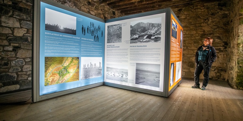 A person in Historic Environment Scotland uniform stands in front of three large, backlit exhibition panels which display text and imagery about Scottish folklore. The panels are in a room with old stone walls, a timber beam ceiling and wooden floor.