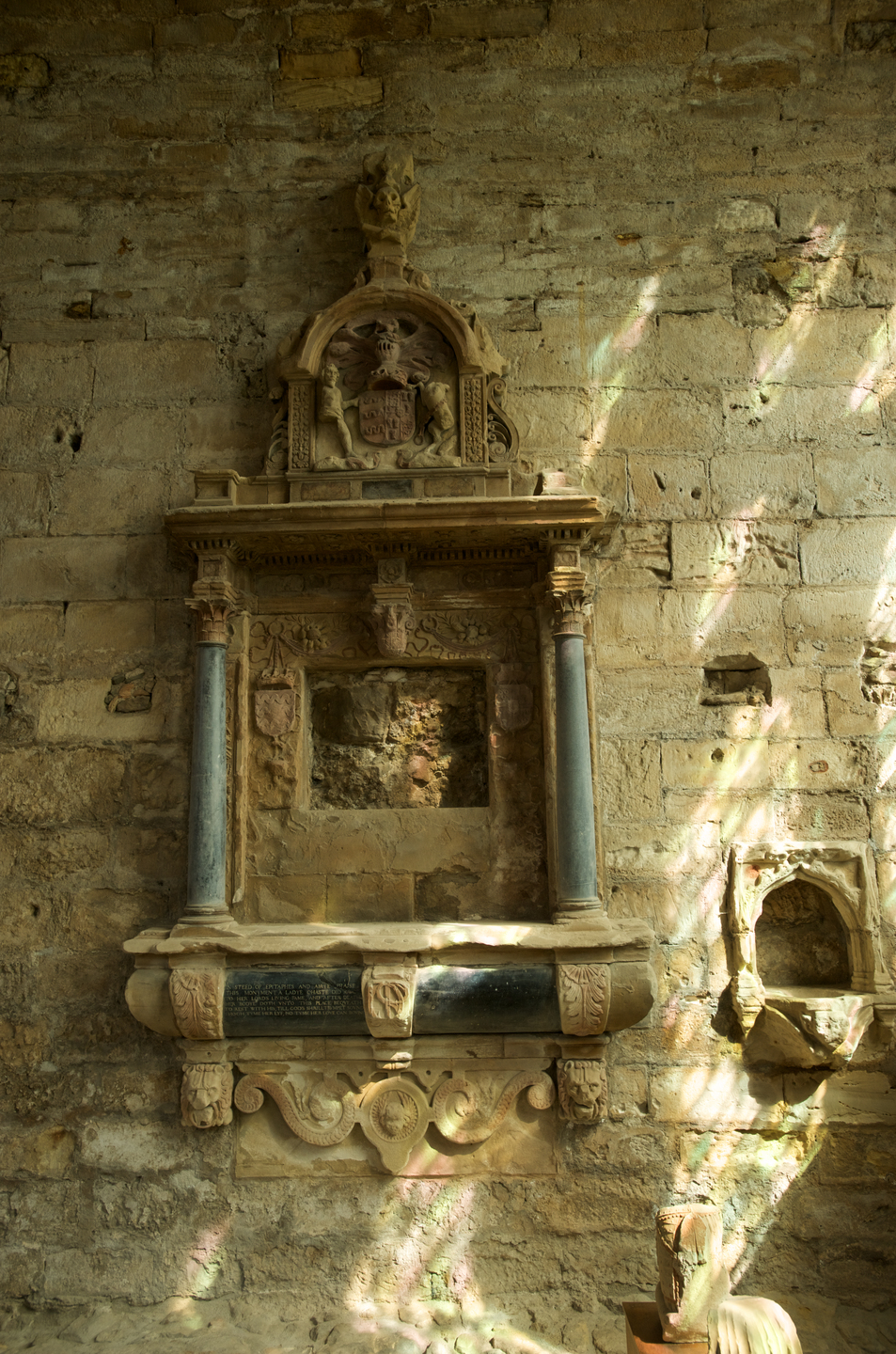 A weathered stone memorial. There are coloured curved columns around the outside, much of the memorial has deteriorated however. There are specks of fragmented light on the wall.