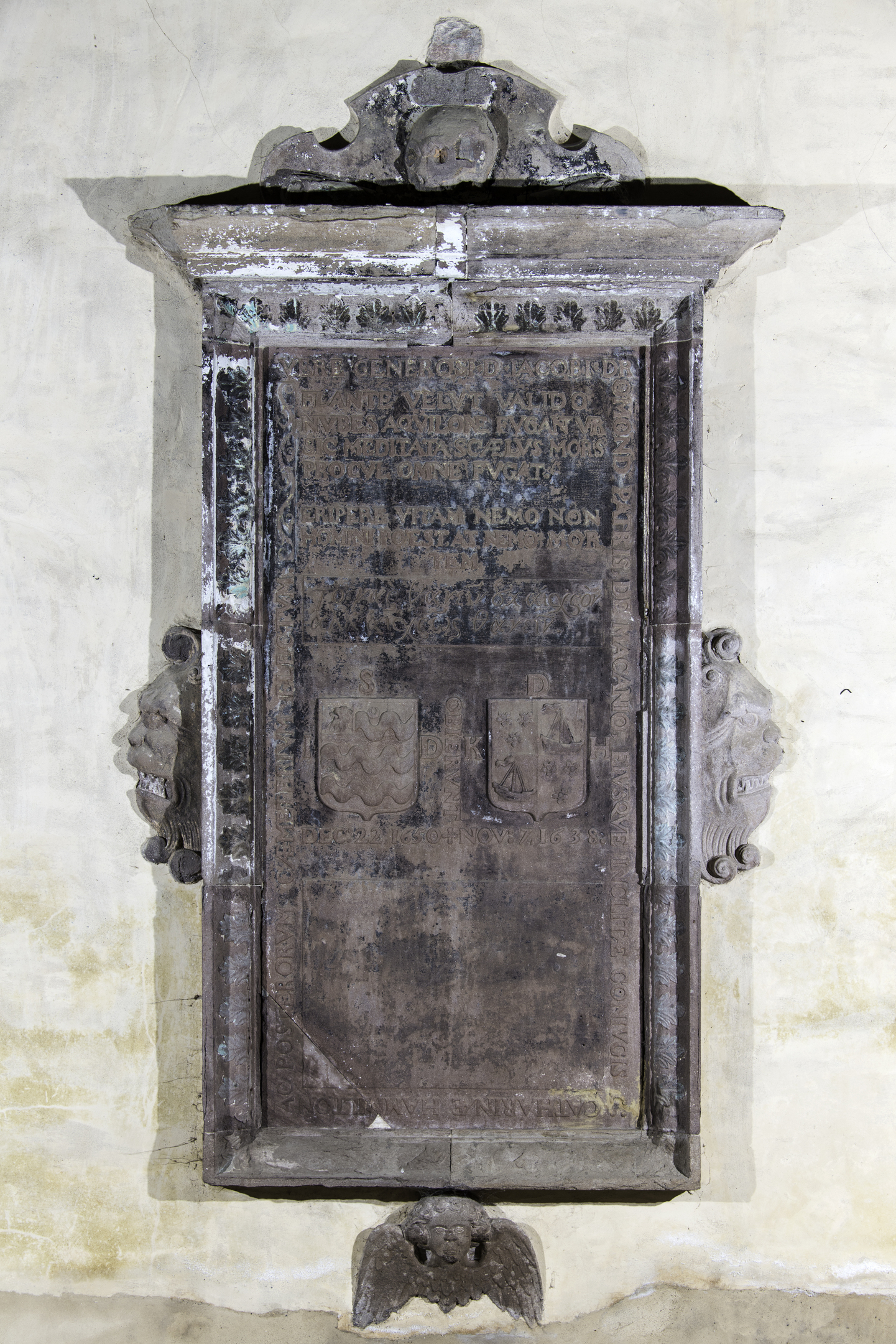 A tall, rectangular stone memorial plaque with heavy weathering, featuring Latin inscriptions, two heraldic shields, and carved lion heads projecting from the sides, topped with an ornate broken pediment and a small winged emblem at the base.