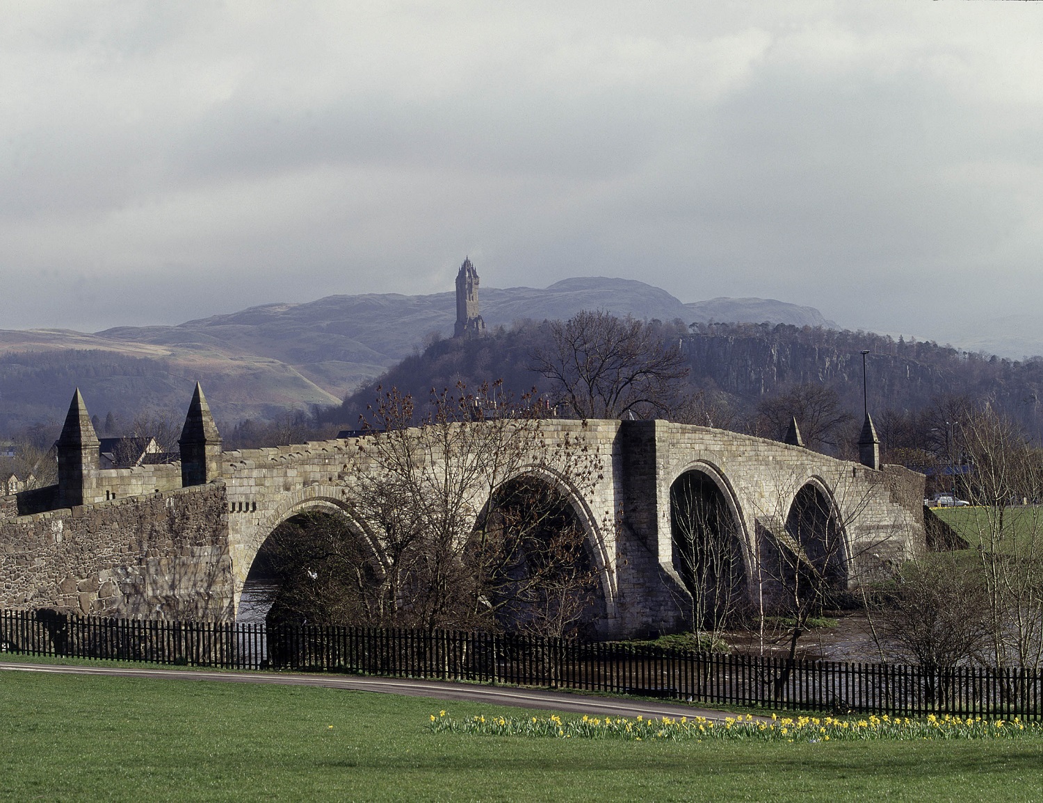 Stirling Old Bridge seen from a grassy riverside, with its series of stone arches stretching across the river. In the background, the Wallace Monument rises on a distant hill beneath an overcast sky. A low wooden fence and spring flowers line the foreground.