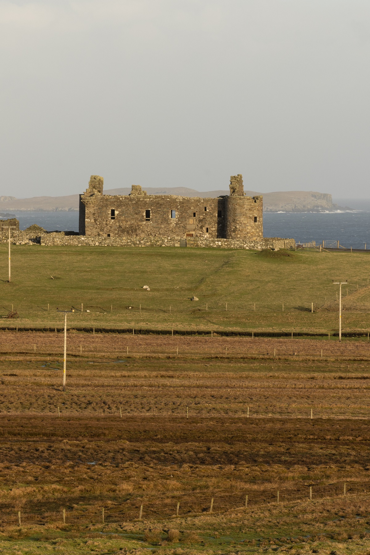 Distant view of Muness Castle, a rectangular stone ruin with two prominent towers, standing on a grassy rise near the coastline. The surrounding landscape is open fields. Beyond the castle, the sea and low rocky islands are visible under a pale sky.