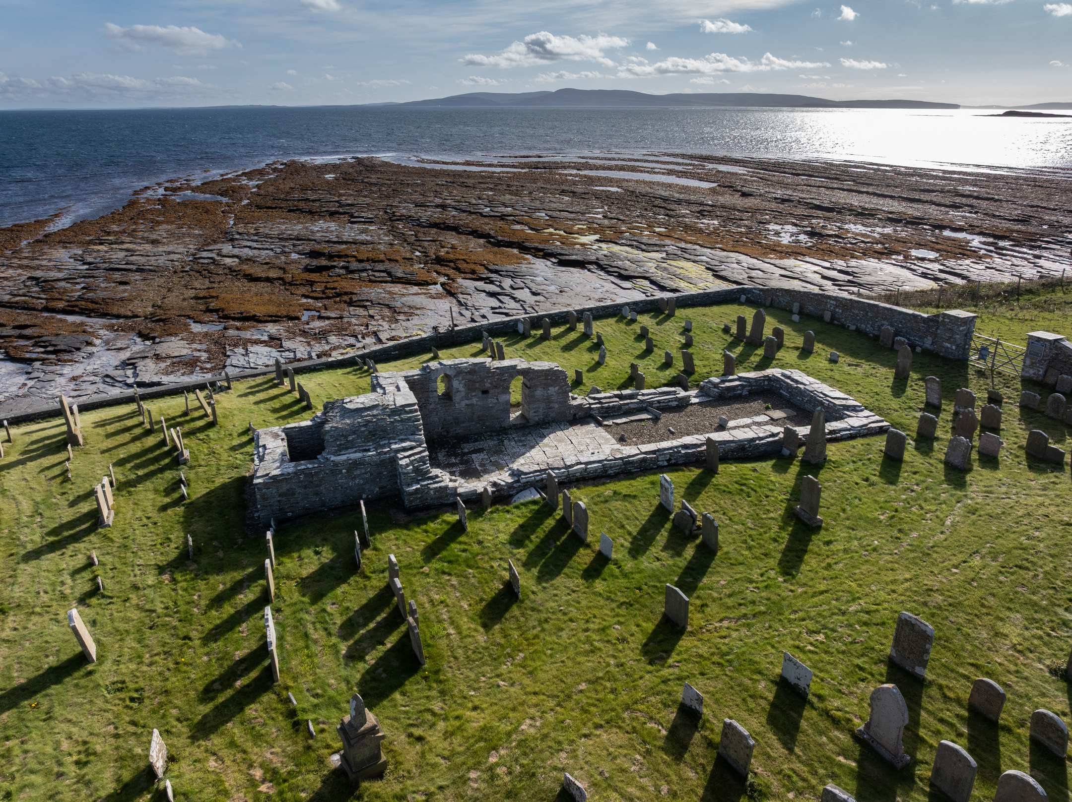 High, wide shot of Westside Church’s ruins and surrounding graveyard situated on a grassy headland overlooking the sea and rocky foreshore.