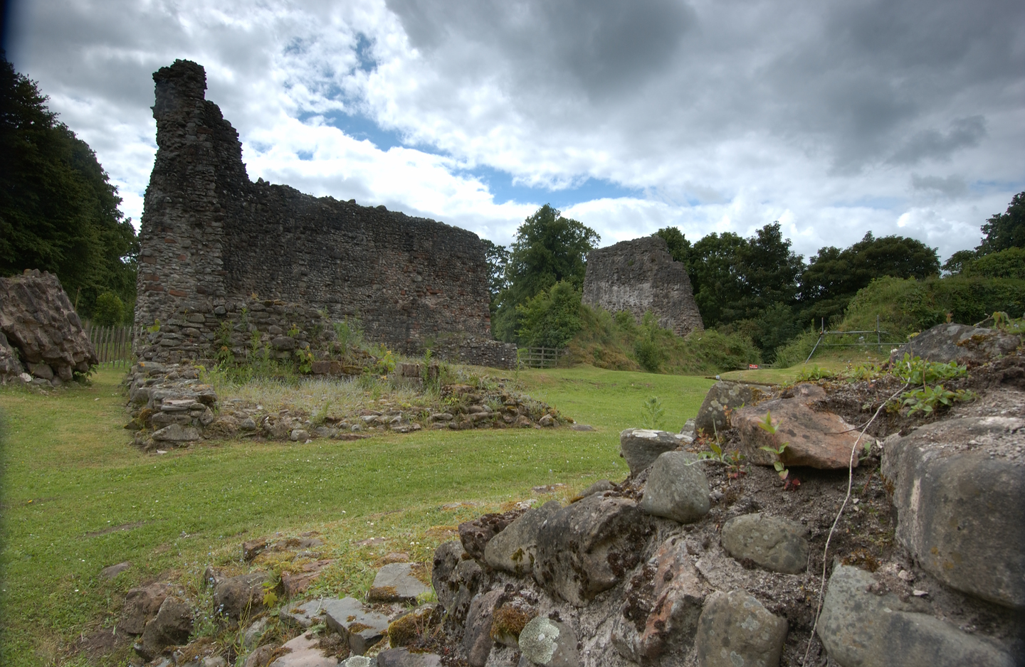 Remains of a historical site, with a close up of parts of a stone wall, with tall stone wall structures in the background which have deteriorated to varying degrees. Trees are in the background. 