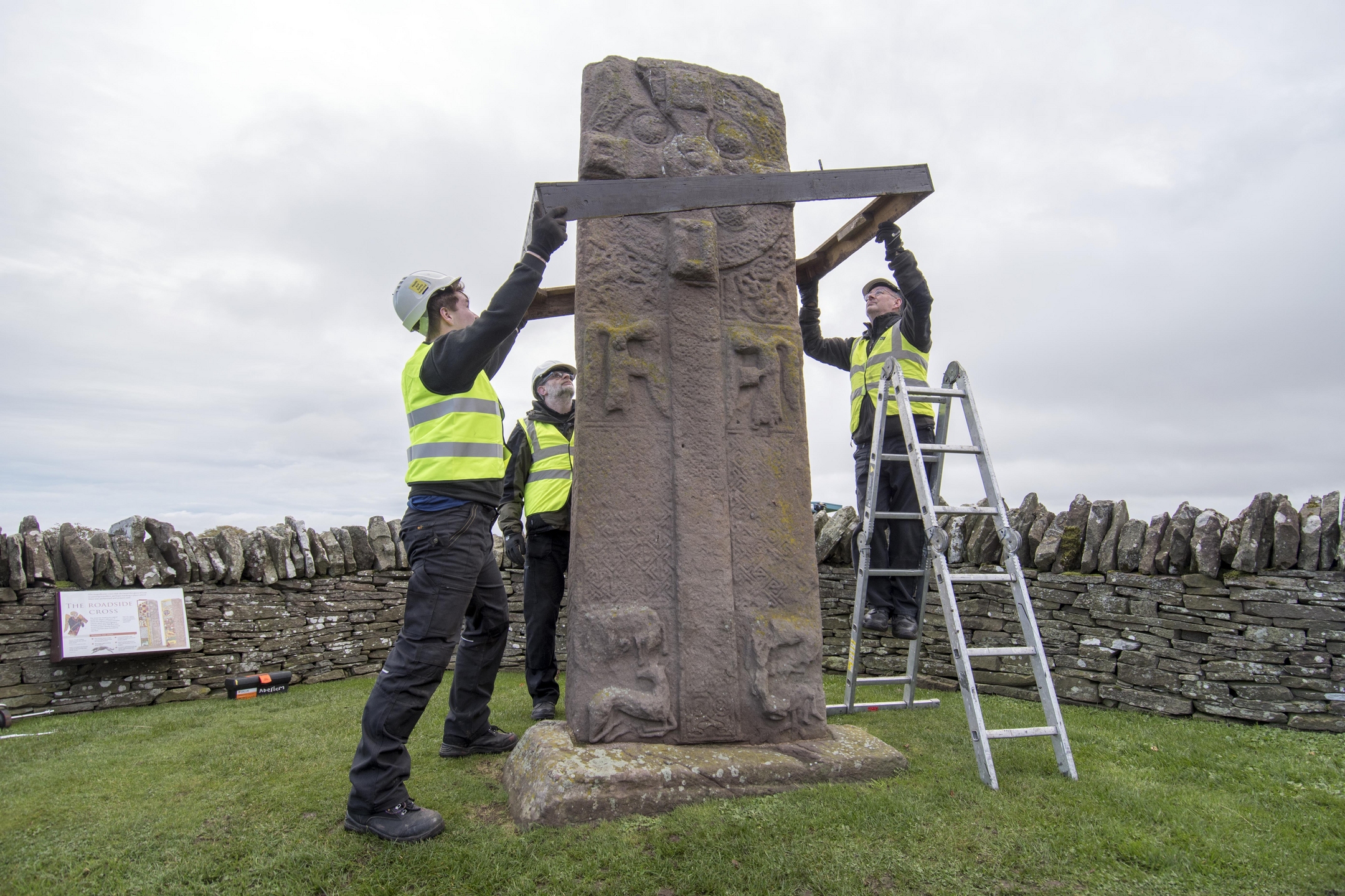 A team of three workers in hi-vis and hard hats begin the process of covering a standing stone to protect it during the winter. They begin by lowering a square wooden frame over the top of the stone.