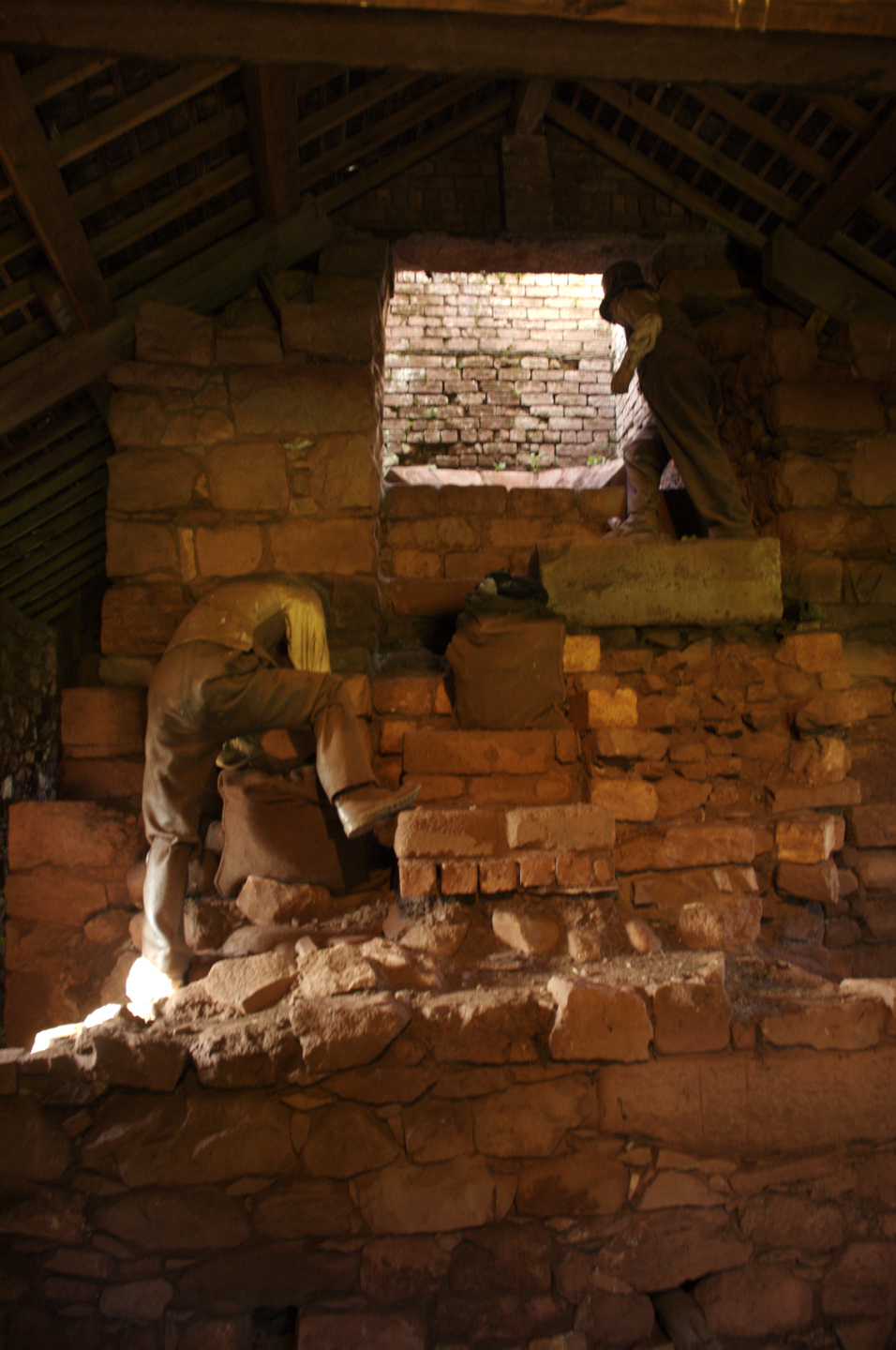 Inside a dim stone building, two life‑sized mannequins dressed as workers stand on stepped stone platforms. One figure bends forward over a bag, while the other stands near a small opening in the wall. The walls and floor are made of rough red stone, and the sloping roof is supported by wooden beams