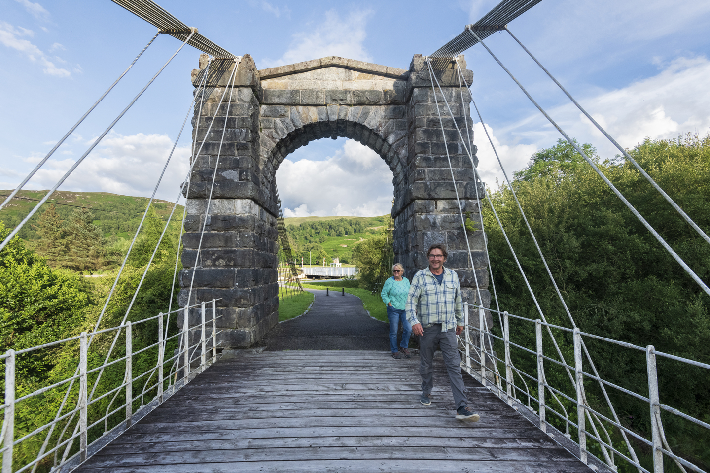 Two people walking across a wooden bridge, with white fencing either side. There is a large stone arch at the entrance of the bridge, and trees either side. 