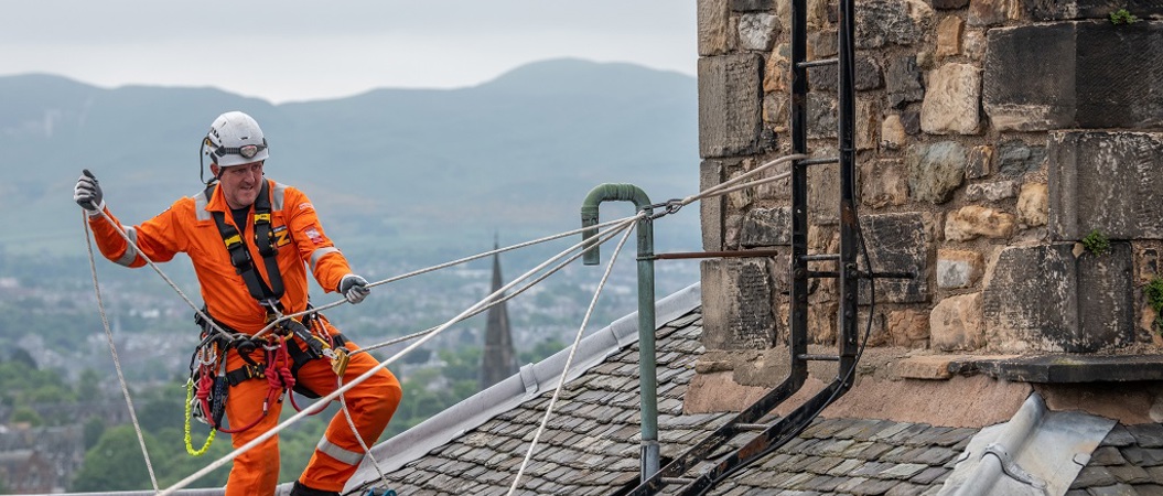 A worker in orange overalls and a hard hat inspects a rooftop at Edinburgh Castle using ropes and a harness.