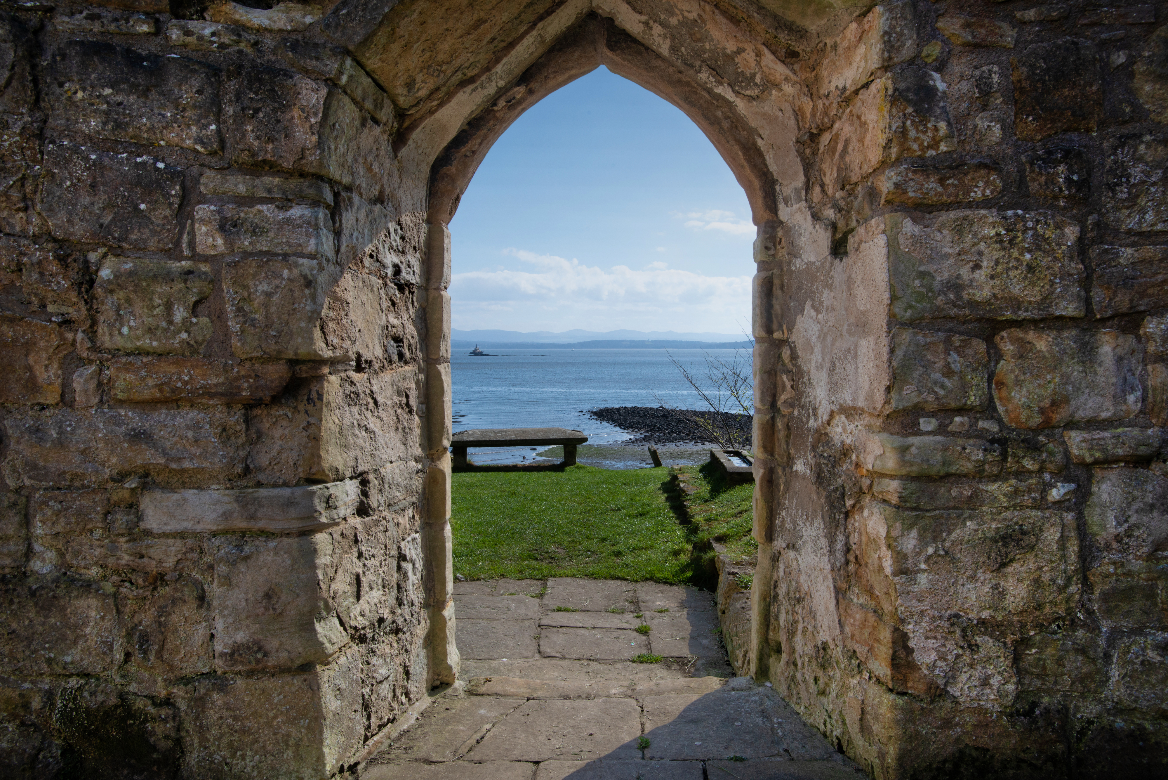A stone archway of the ruined kirk framing a view of the sea, with a bench and grassy area leading to the shoreline beyond.