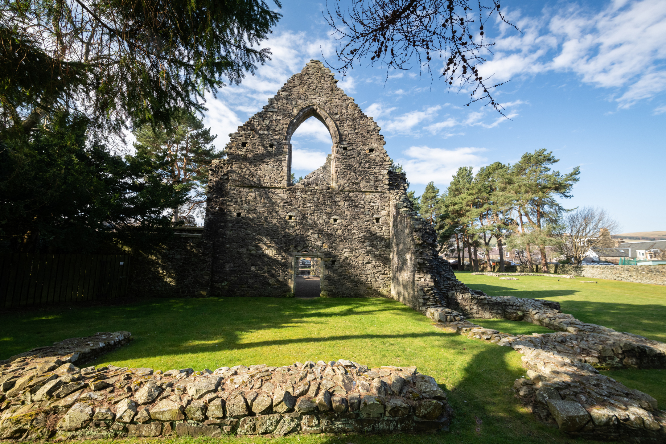 The ruins of the Cross Kirk ruins in Peebles. The stonework is weathered and partially collapsed on the sides. Sunlight casts shadows across the grassy area in front.