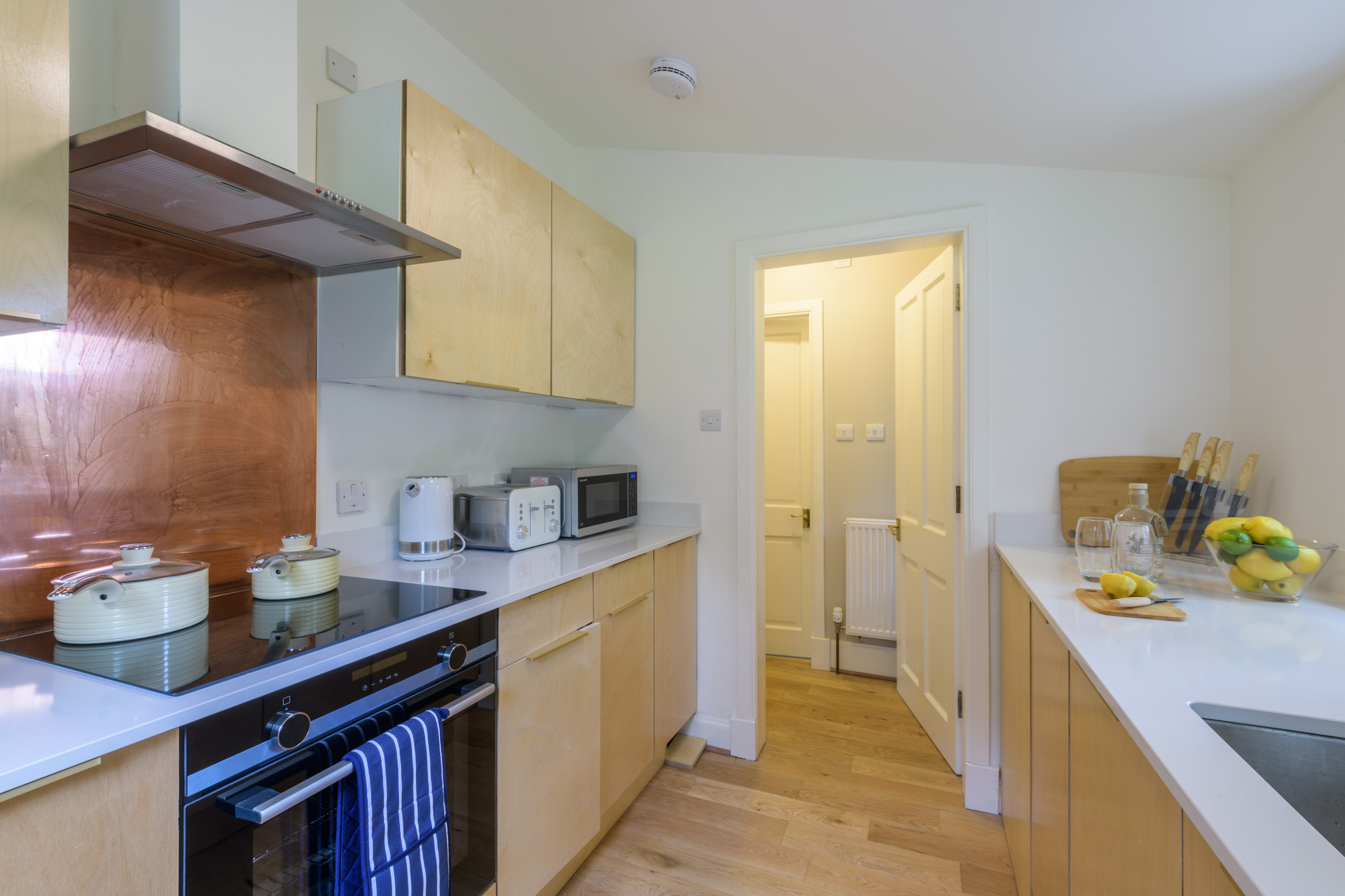 A galley kitchen with wooden units, white work tops, white walls and a wooden floor