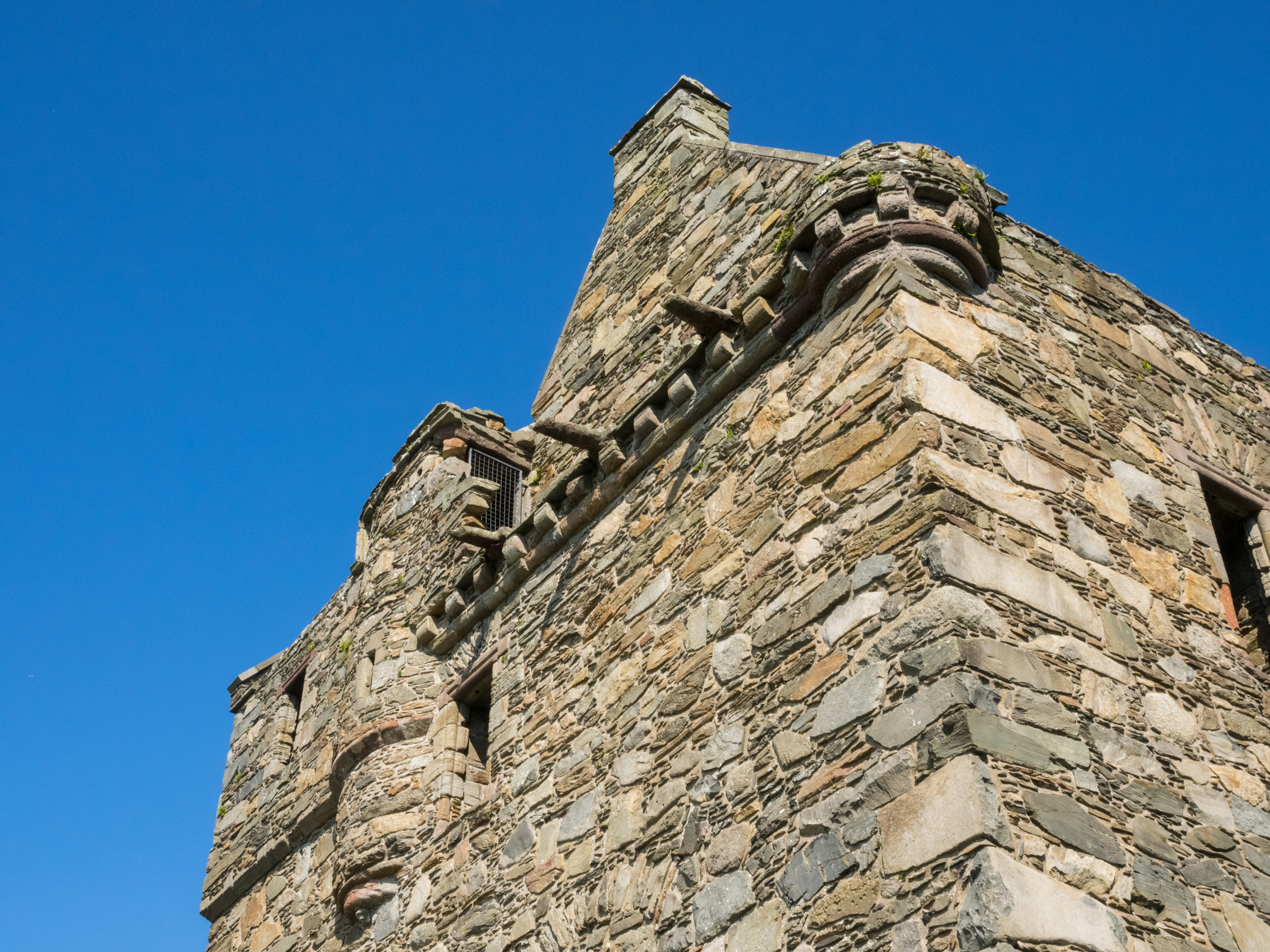A close, low‑angle view of the upper section of Carsluith Castle, highlighting its stonework, corbelled parapet, and gable roofs set against a clear blue sky.