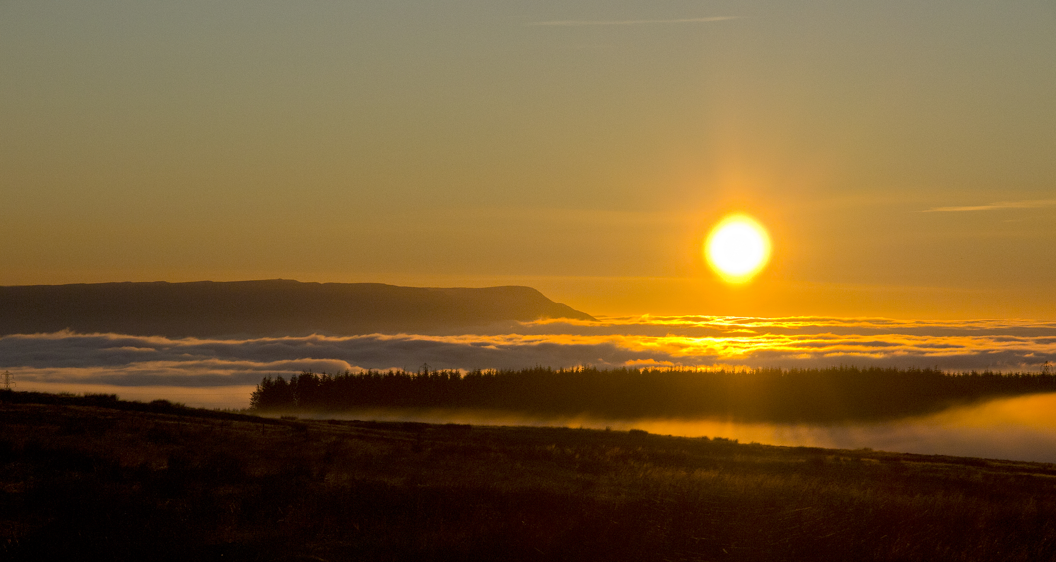 Golden sunrise above a blanket of low cloud, with the sun hanging over distant hills and dark foreground moorland silhouetted against the warm sky.