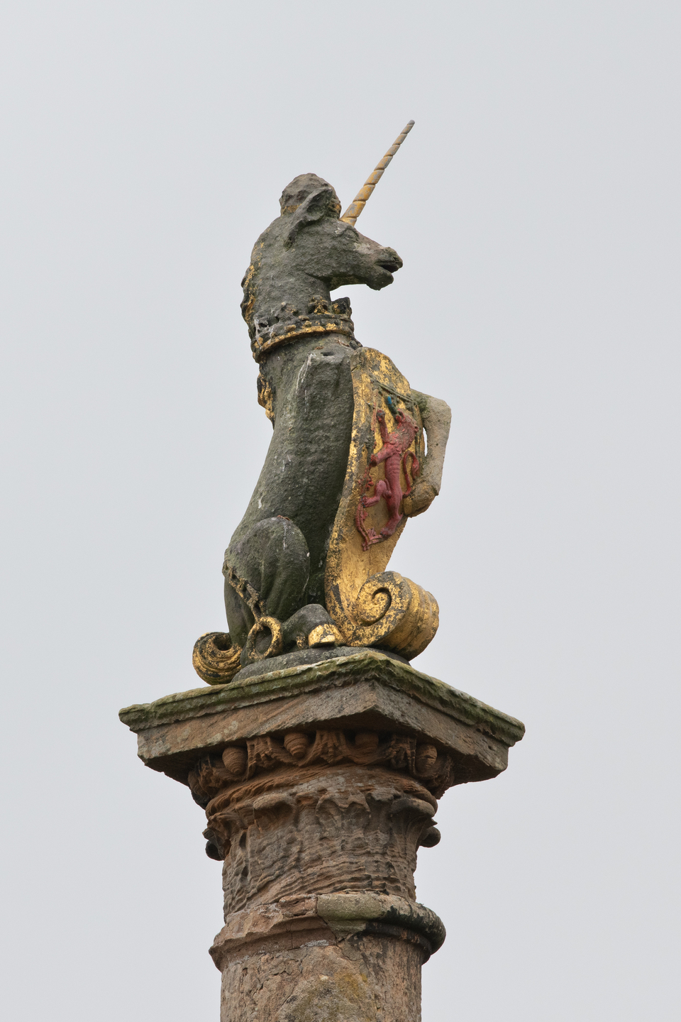 The top of a statue, where a stone unicorn sits. Its body is facing away. There are markings of gold on it, particularly it's front. The mouth of the unicorn is slightly open. 