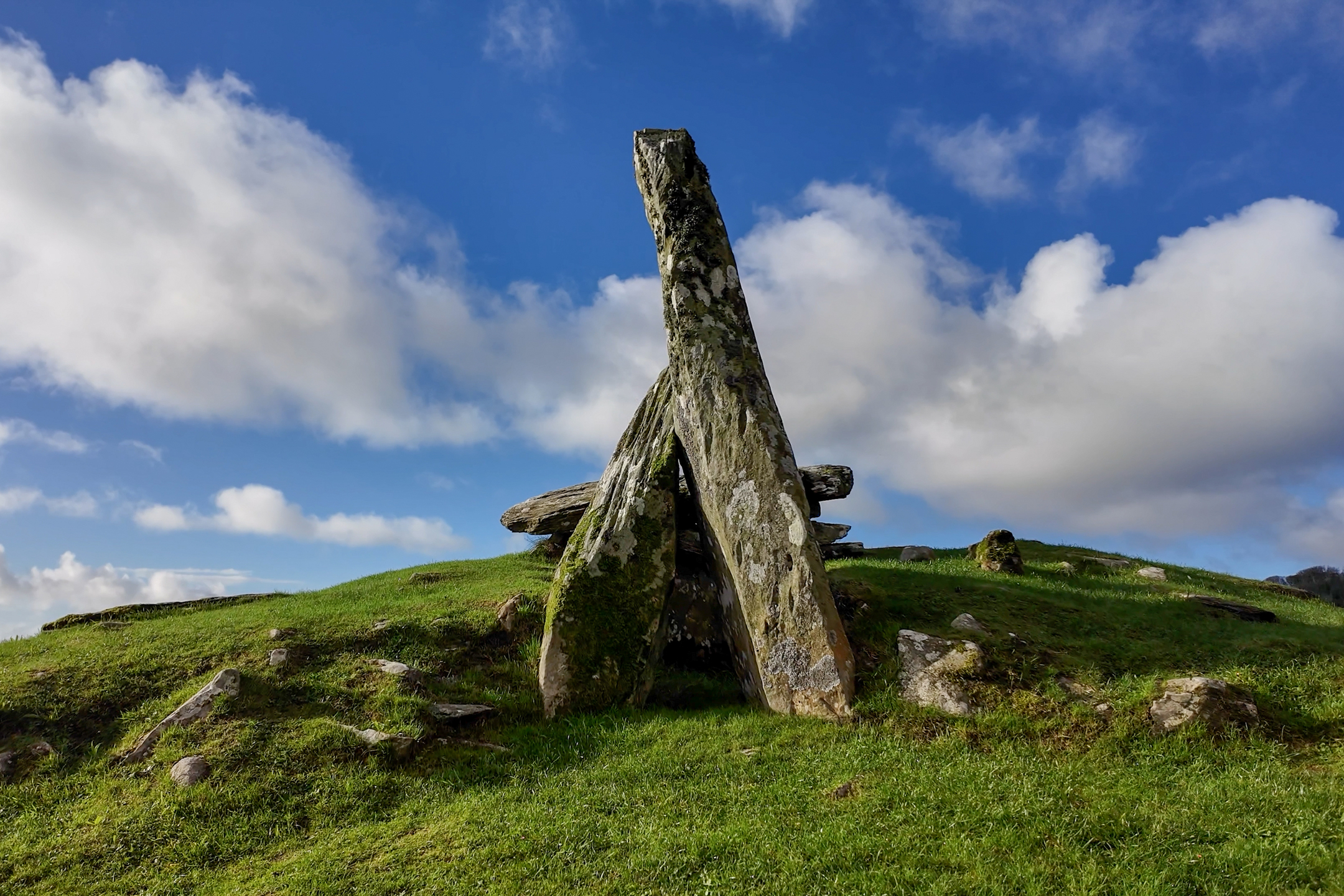 Stone burial monument formed by tall, leaning slabs creating a triangular entrance, set on a grassy hill under a blue sky with scattered clouds.