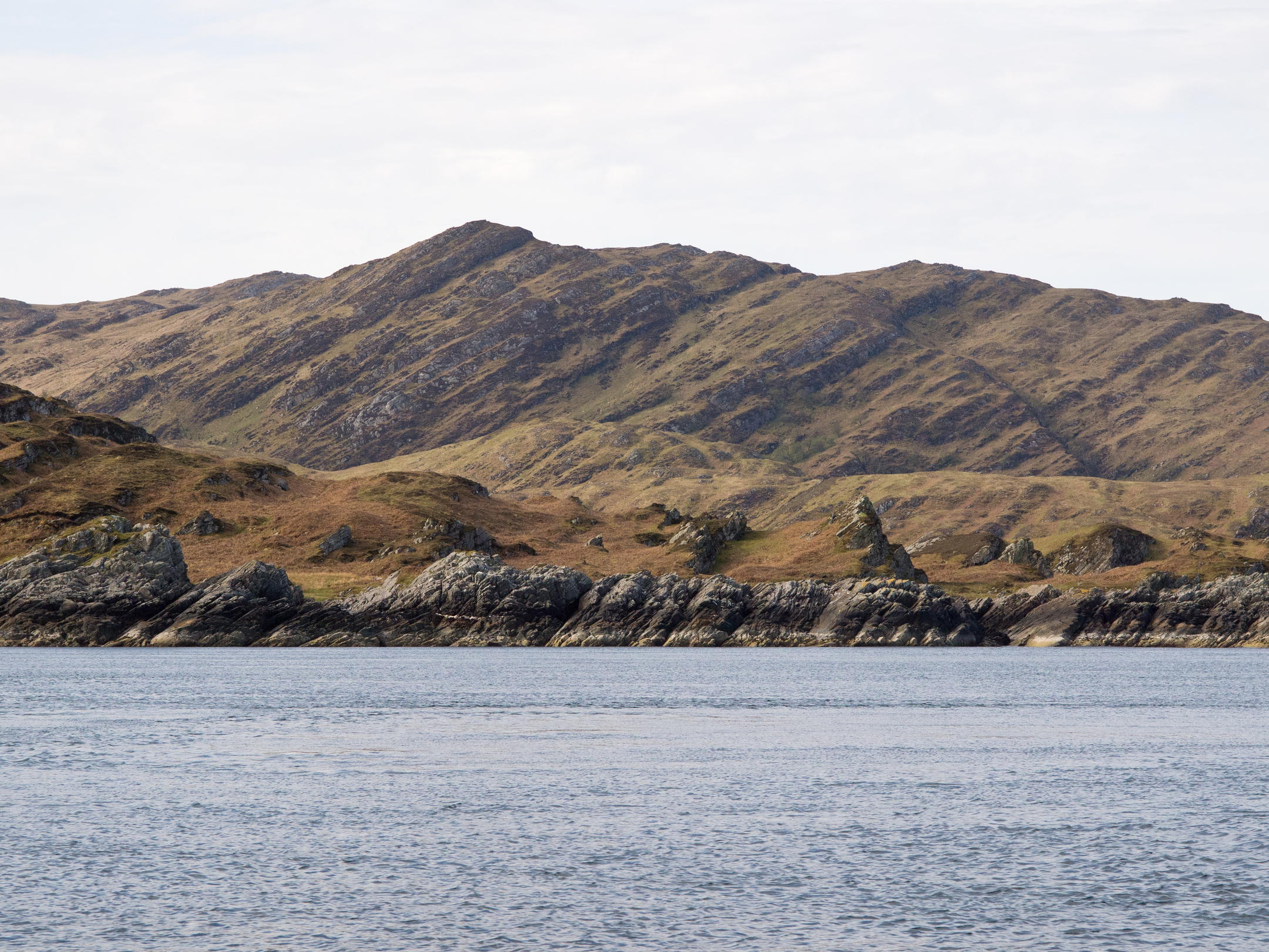 Rocky coastline with jagged outcrops and calm blue water, backed by rugged hills under a partly cloudy sky.