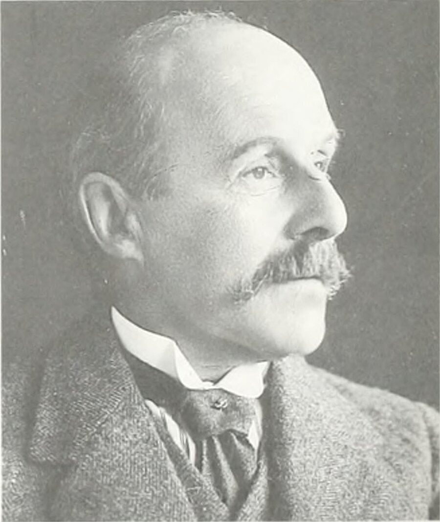 Black and white portrait photo of a person looking to the right of camera. They are wearing an old-style suit and thin tie, and have bushy hair around their ears.